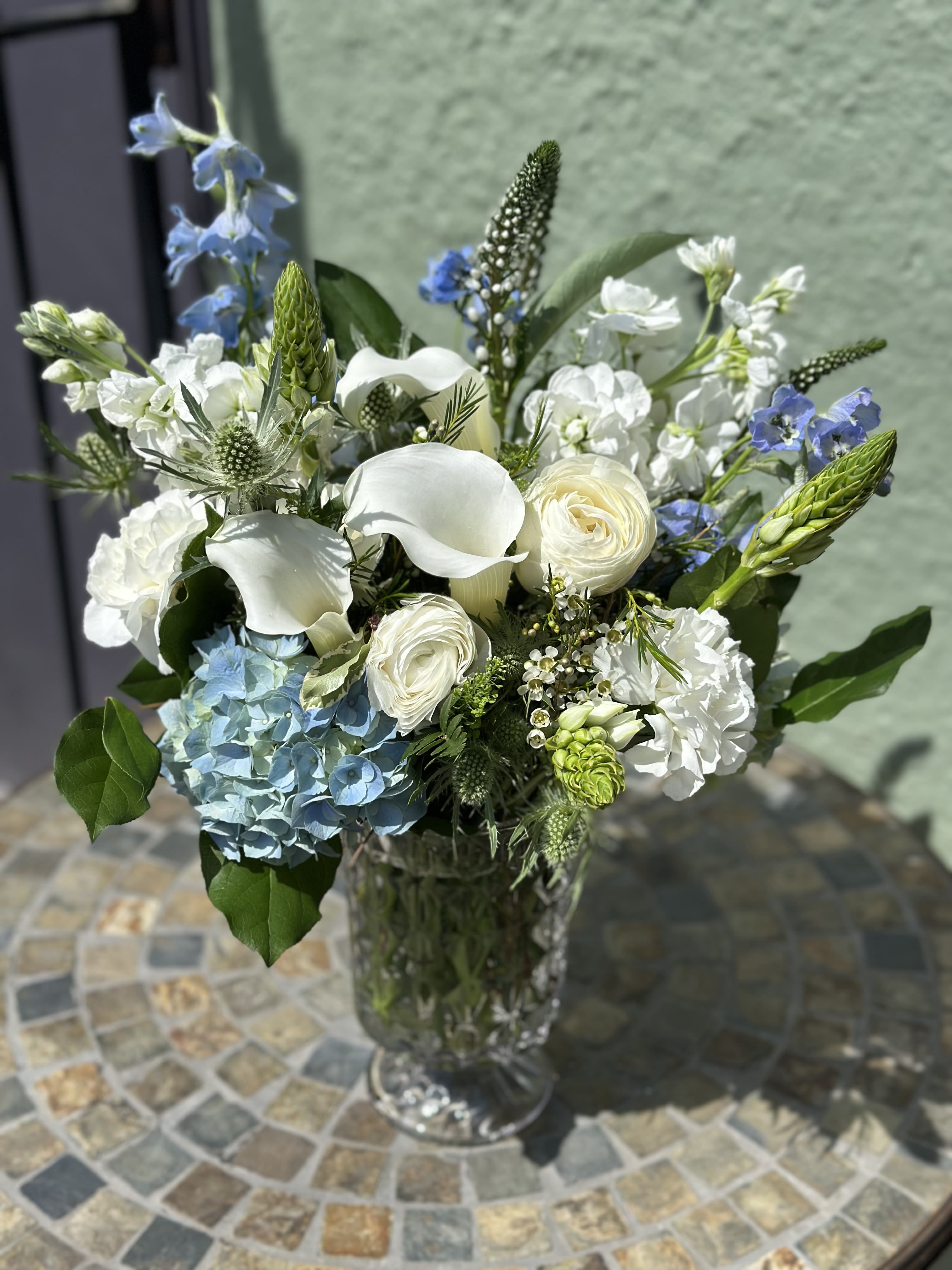 Arrangement of white calla lilies, roses, and blue hydrangea in a tall clear glass vase