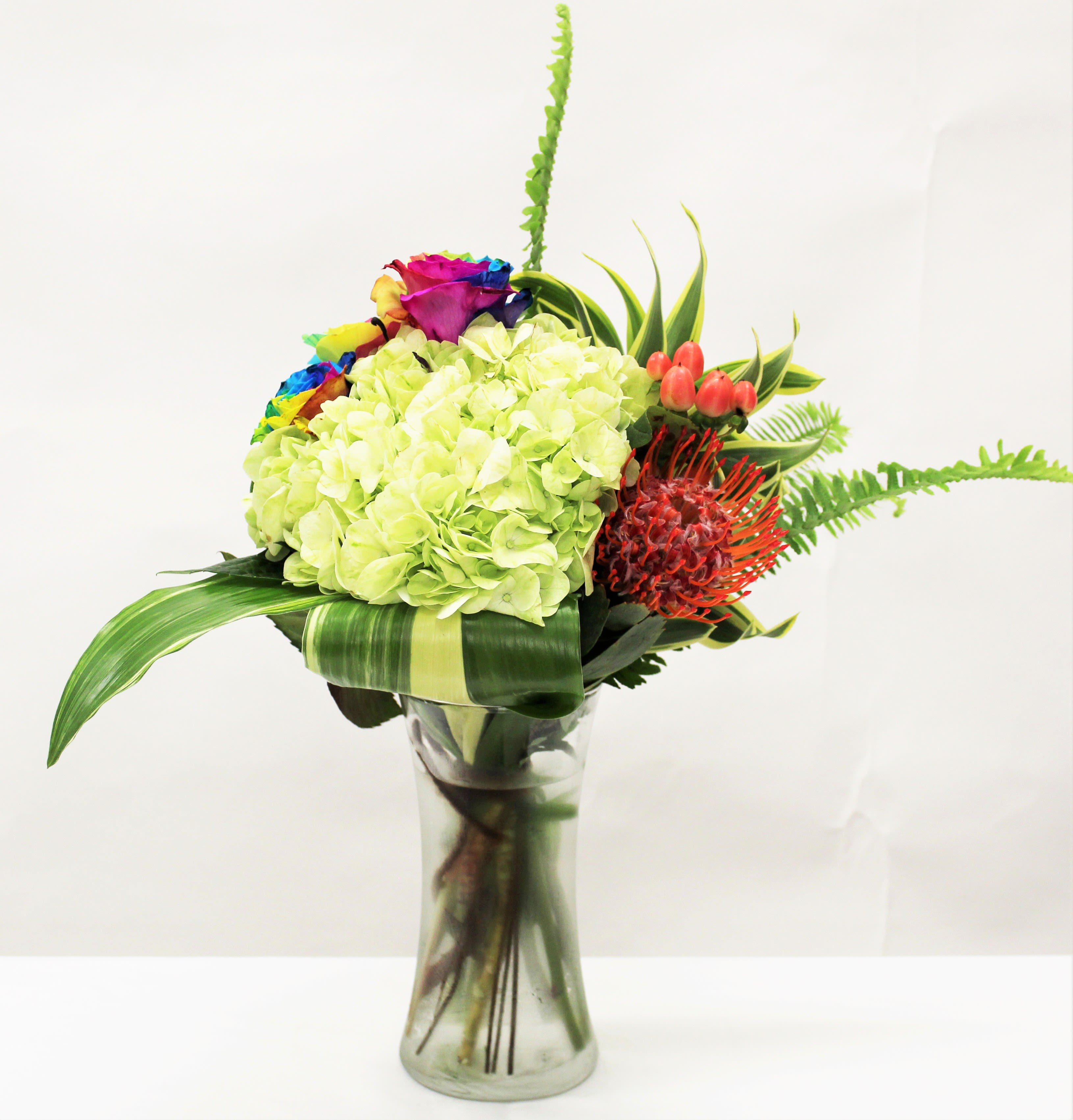 Modern bouquet of green hydrangeas, rainbow roses, and orange protea in a clear glass vase.