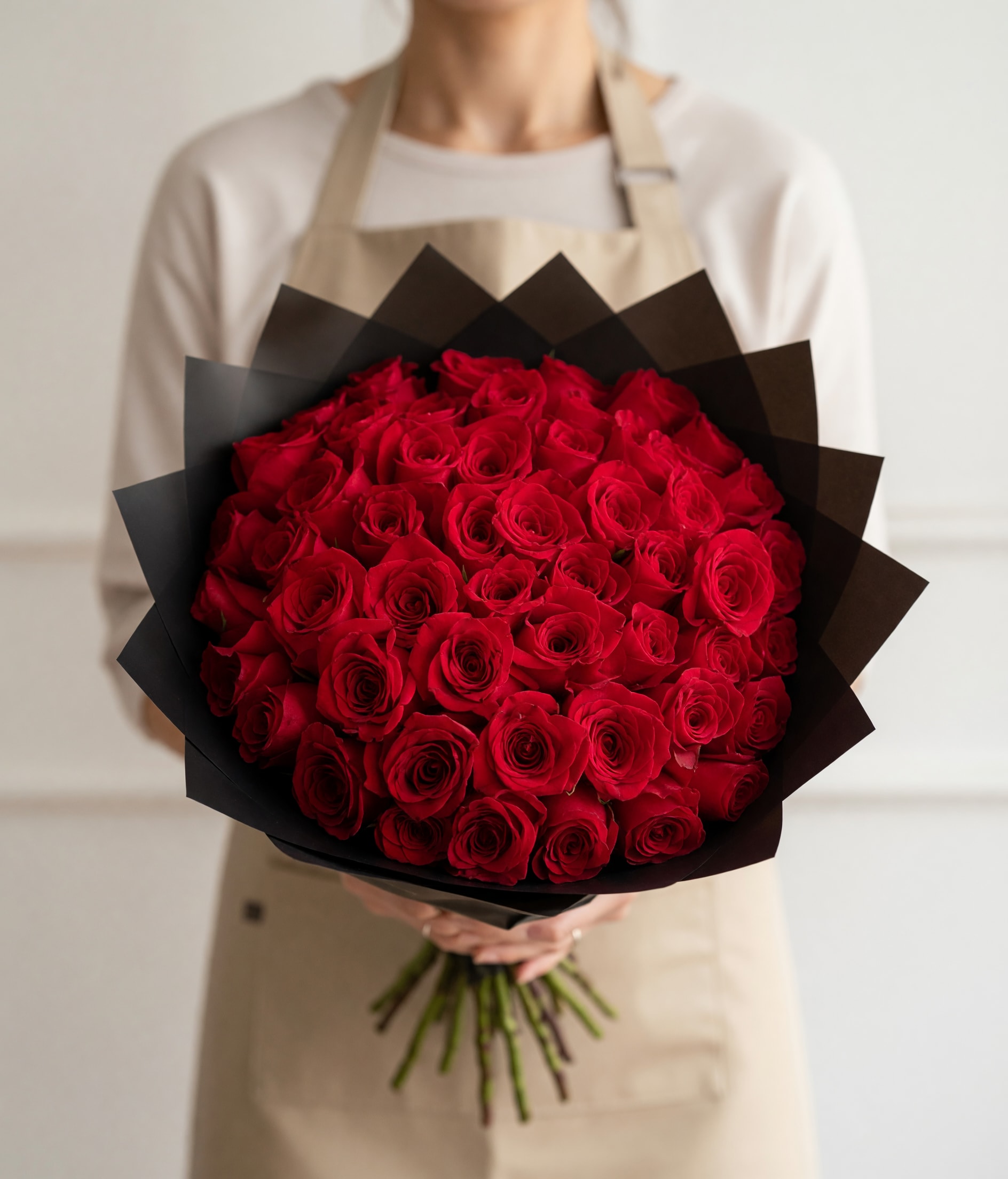 Roses  (50, 75, 100) Buchon - A bold, dome-shaped bouquet of tightly packed red roses that reads as classic romance with a modern edge - perfect as an anniversary roses bouquet or a Father's Day surprise with dramatic flair. The rich crimson petals create a full, luxurious presence that makes the person you're thinking of feel deeply celebrated; the striking black paper wrap gives the arrangement a contemporary, gift-ready polish ideal for Valentines, birthdays, or any occasion meant to say "I love you" or "congratulations."  Each arrangement is composed solely of red roses arranged in a dense, rounded form so every bloom faces outward for maximum visual impact. The angular black paper wrap frames the dome and heightens color contrast, lending a formal, elegant silhouette that also suits sympathy, memorial, or corporate gifting. Available in 50, 75, or 100 roses to match the moment - from meaningful gestures to grand declarations - and designed to travel well while arriving ready to display. Simple care: keep in a cool spot, refresh water daily, and trim stems for longer vase life so the recipient can enjoy the roses for days.