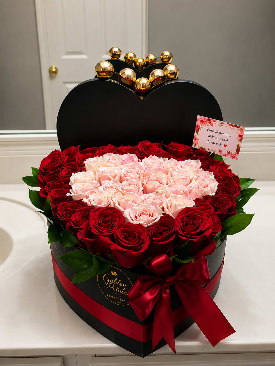 Heart-shaped box of red and blush roses with a red ribbon and gold topper
