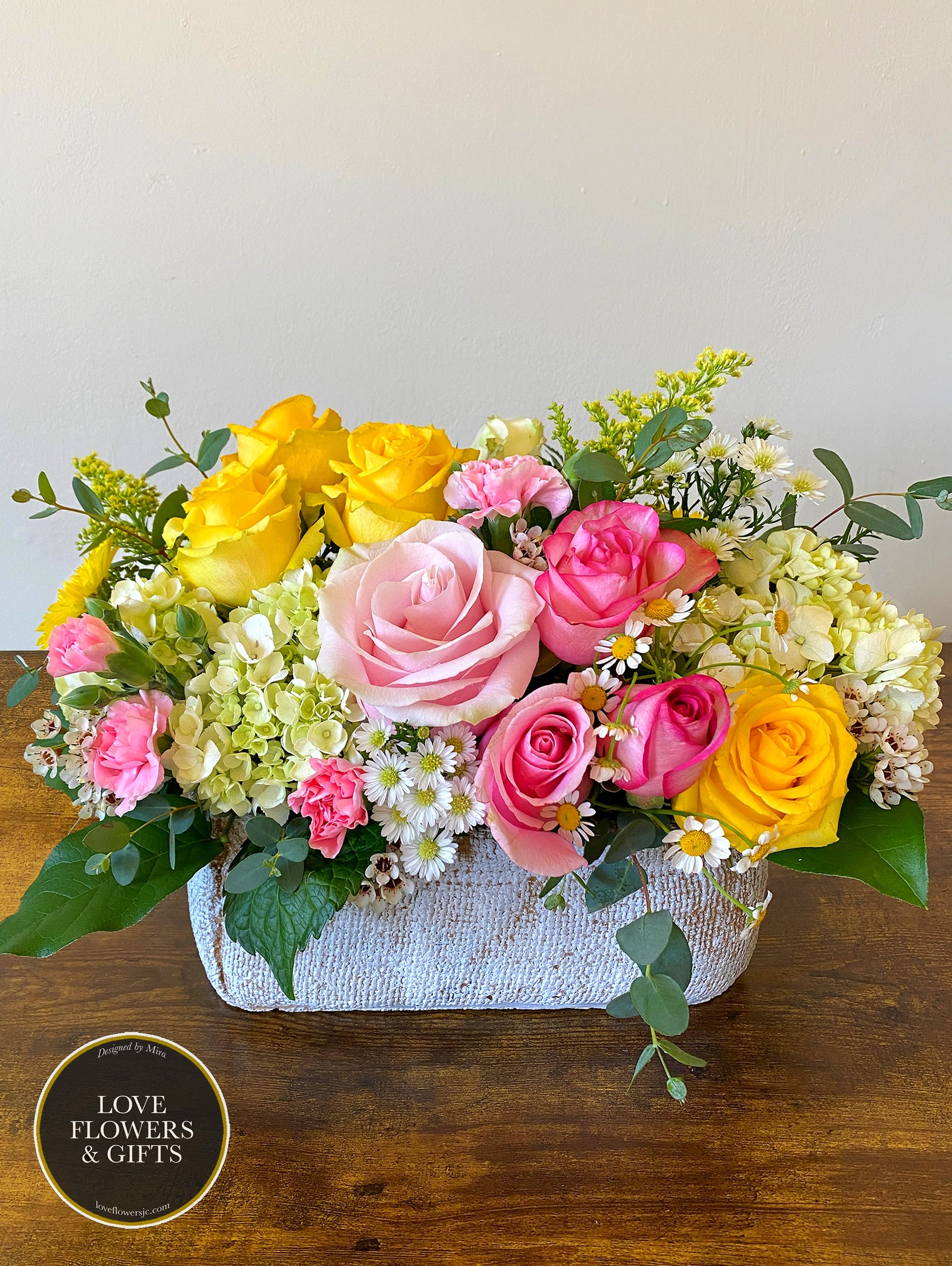 Low arrangement of pink and yellow roses with hydrangeas and daisies in a rectangular white container
