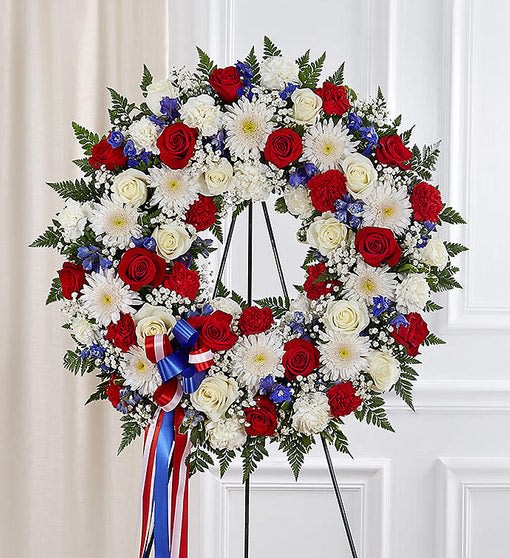 Standing wreath of red roses, white mums, and blue flowers with a patriotic ribbon on an easel.