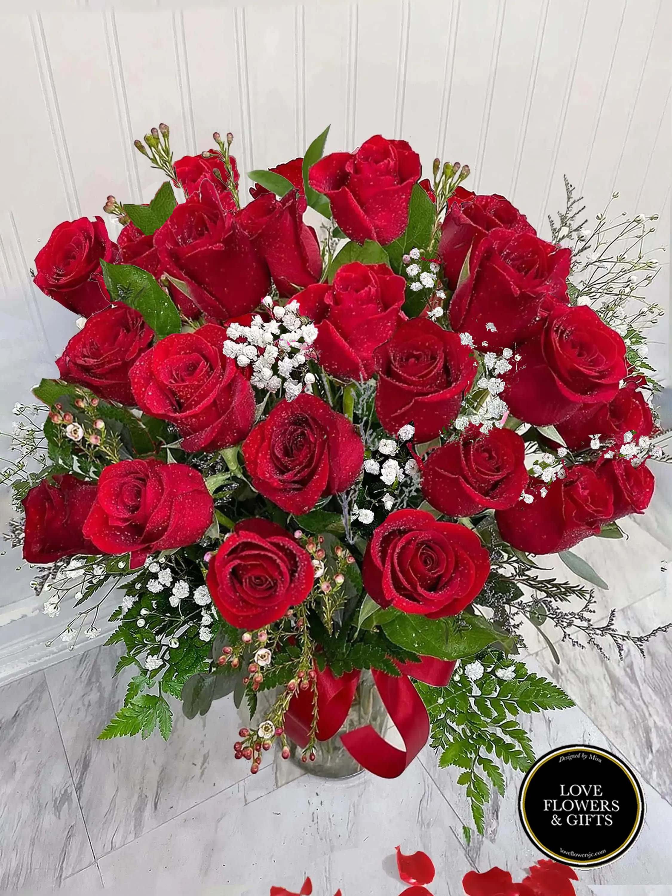 Tall bouquet of red roses with white filler flowers in a clear glass vase with a red ribbon