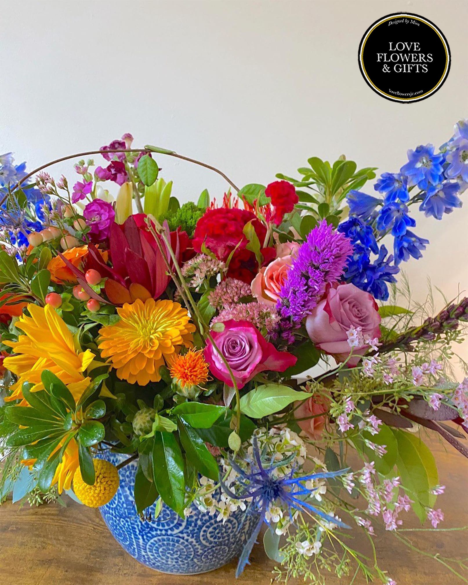 Colorful mixed flower arrangement with roses, delphinium, and daisies in a blue patterned ceramic pot.