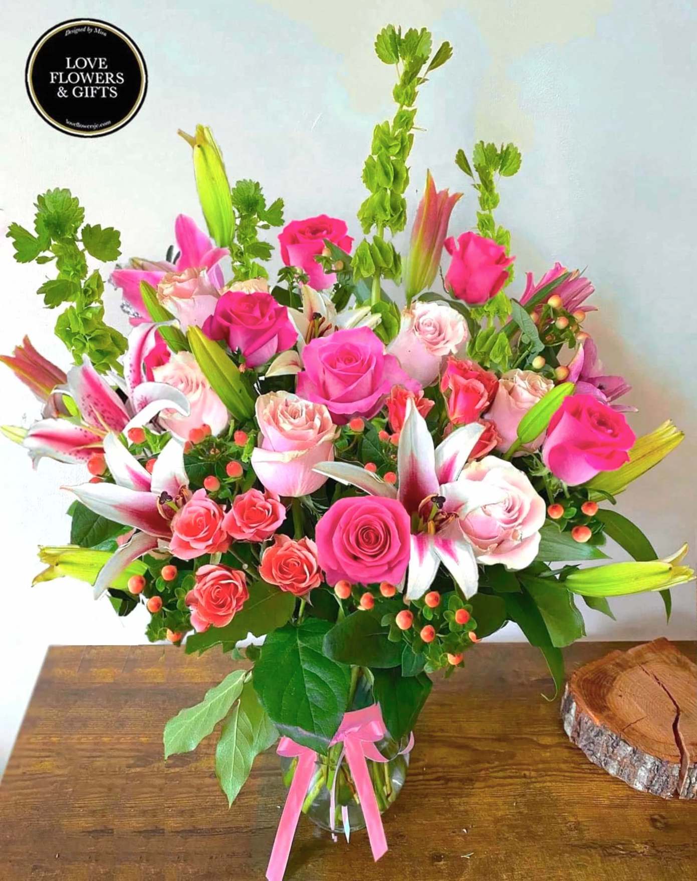 Tall mixed bouquet of pink roses and white-pink lilies in a clear glass vase with a pink ribbon.