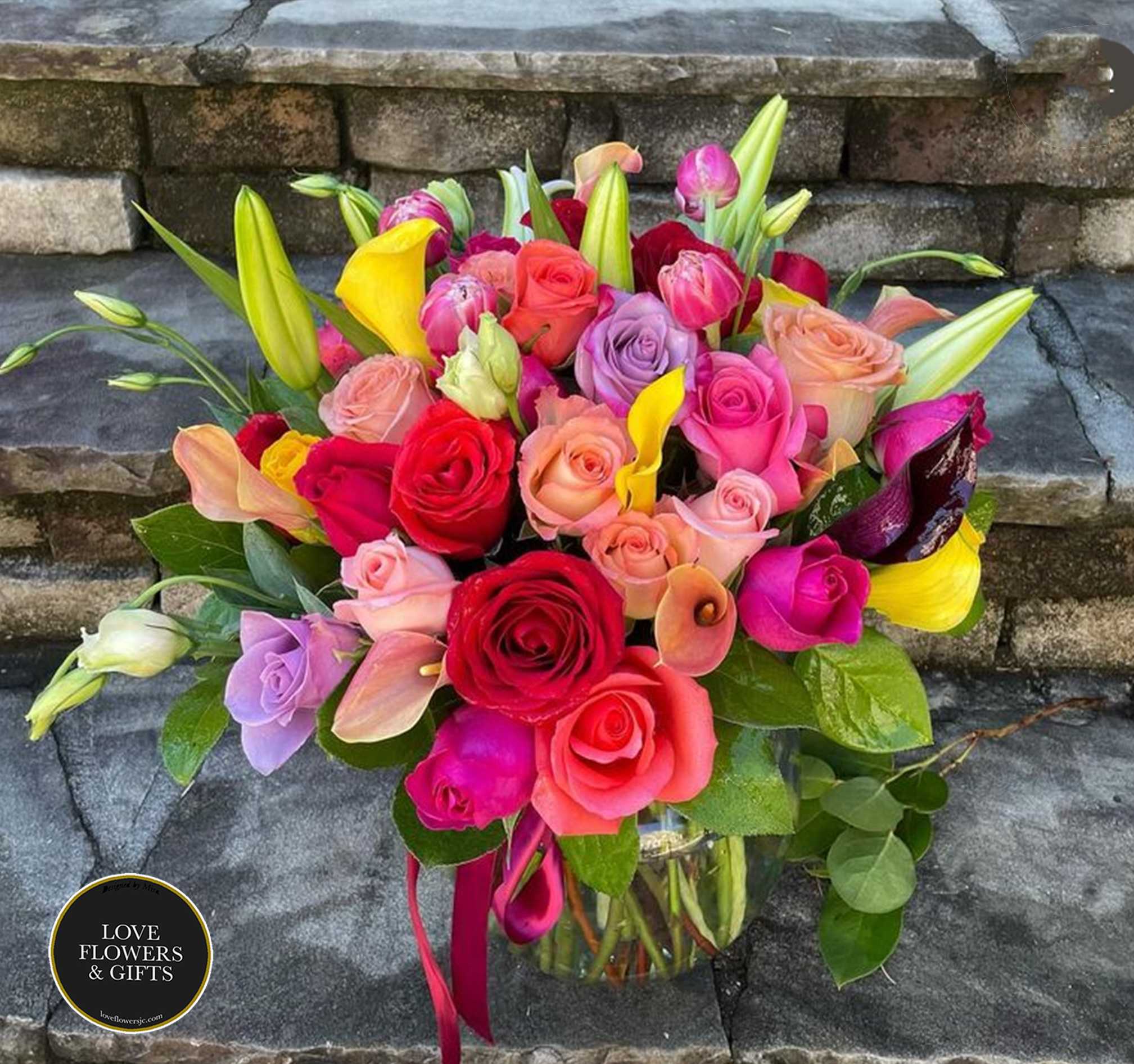 Colorful bouquet of roses, calla lilies, and lilies in a clear glass vase on stone steps
