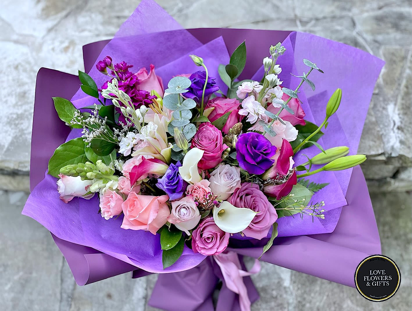 Hand-tied bouquet of pink and purple flowers with white calla lilies wrapped in purple paper.