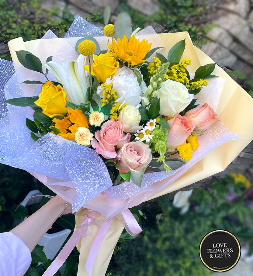 Hand-tied bouquet with yellow sunflowers, roses, white lilies, and pastel roses wrapped in yellow and lilac paper.