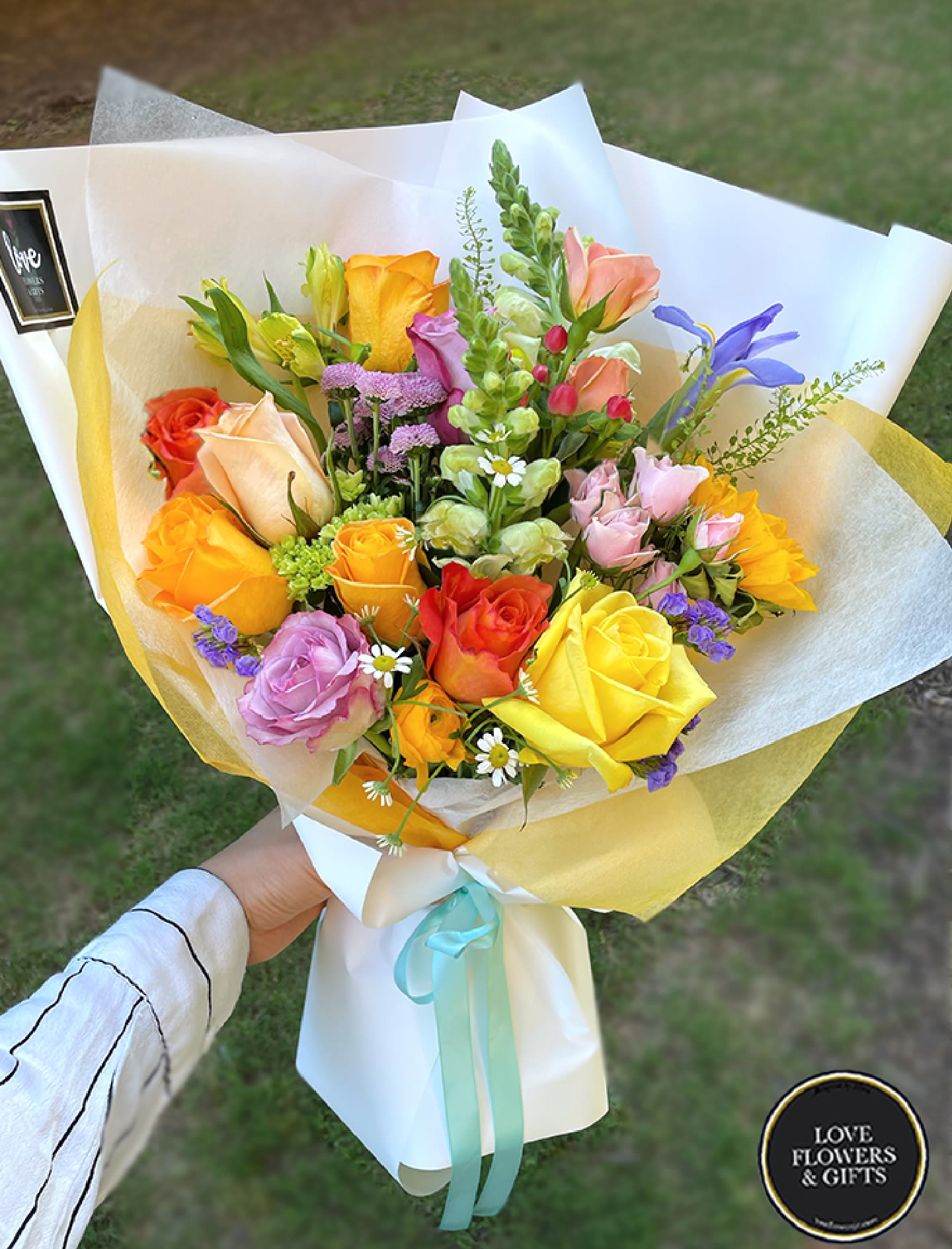 Hand-tied bouquet of yellow, orange, and pink roses with mixed blooms in white wrap.