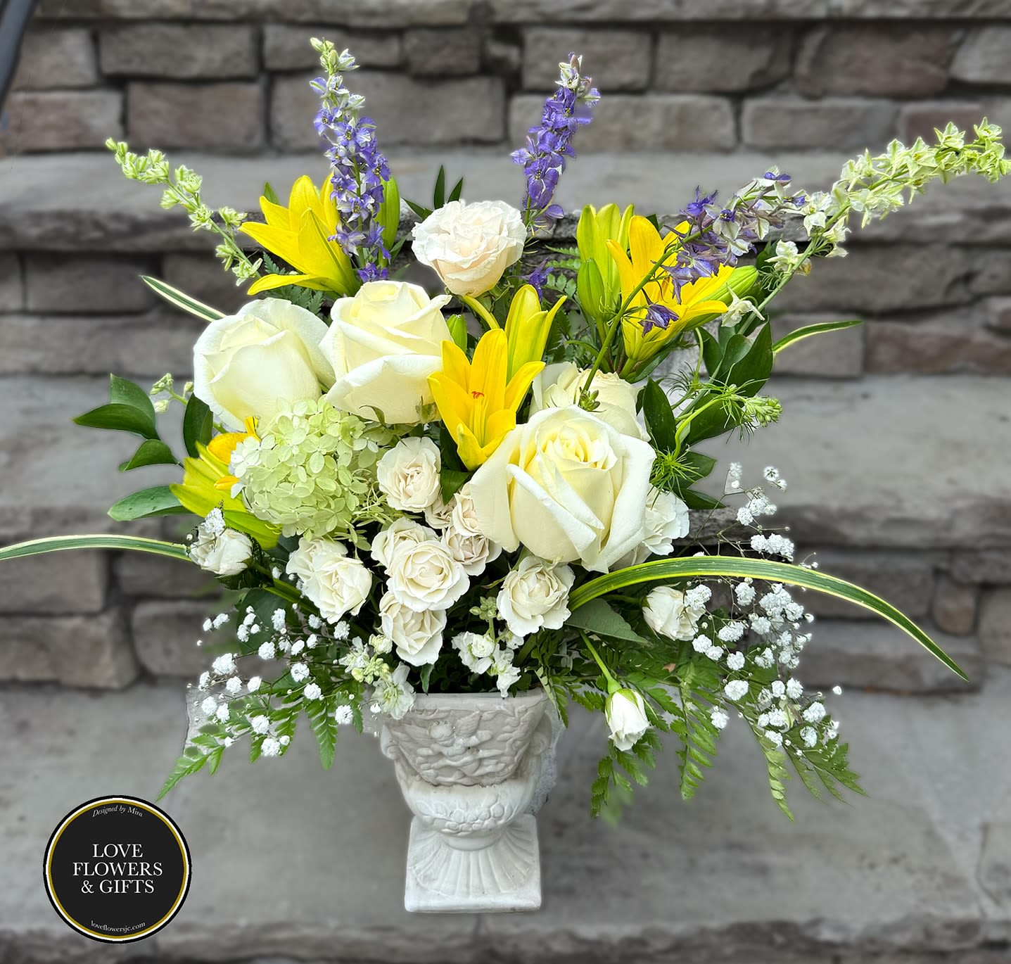 Tall white and yellow flower arrangement with roses and lilies in a white pedestal urn