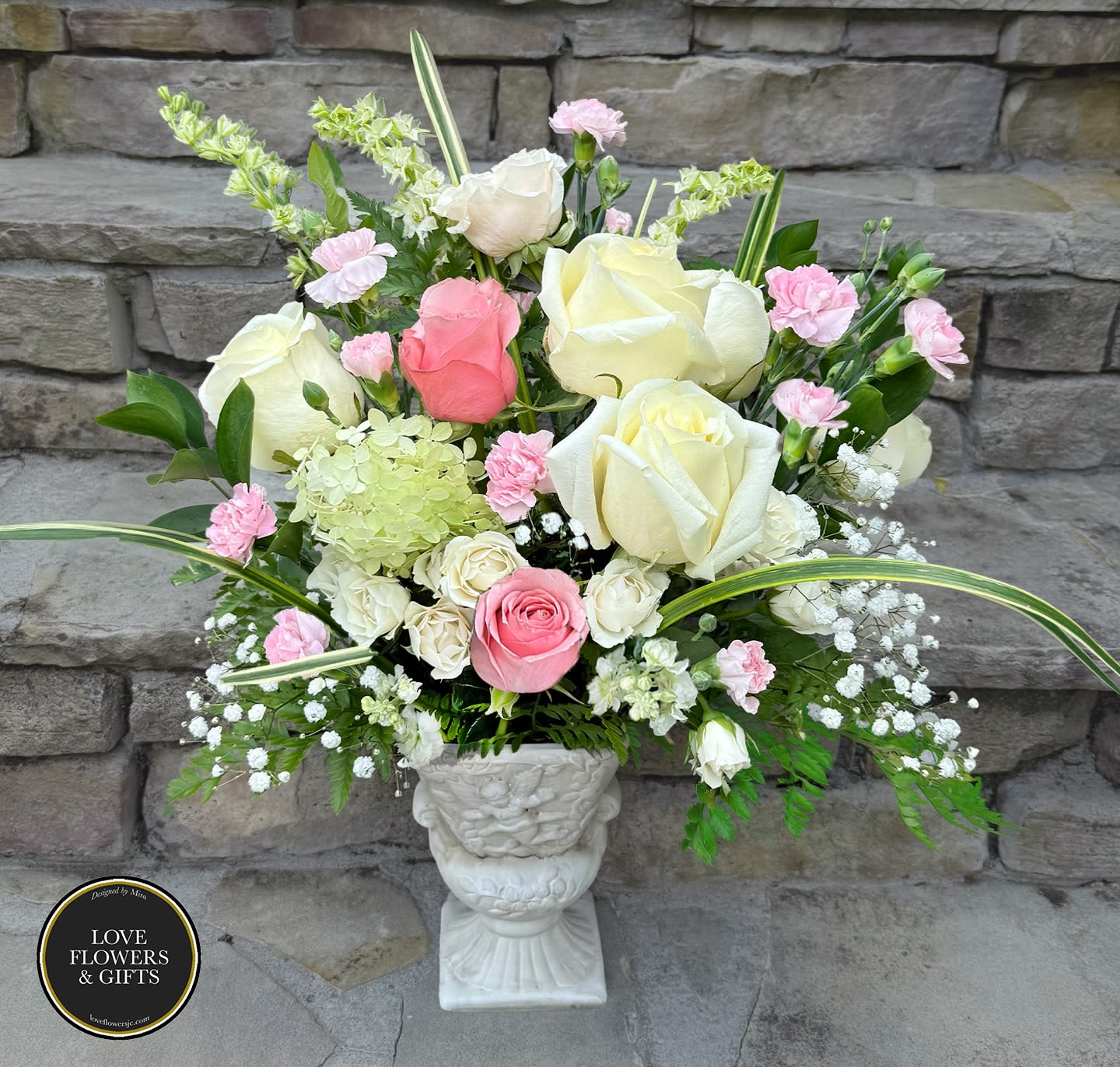 Tall arrangement of white and pink roses with pink carnations and green hydrangea in a white urn vase.