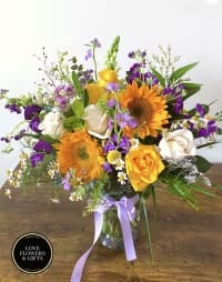 Mixed arrangement of yellow sunflowers, roses, and purple flowers in a clear vase with a lavender bow