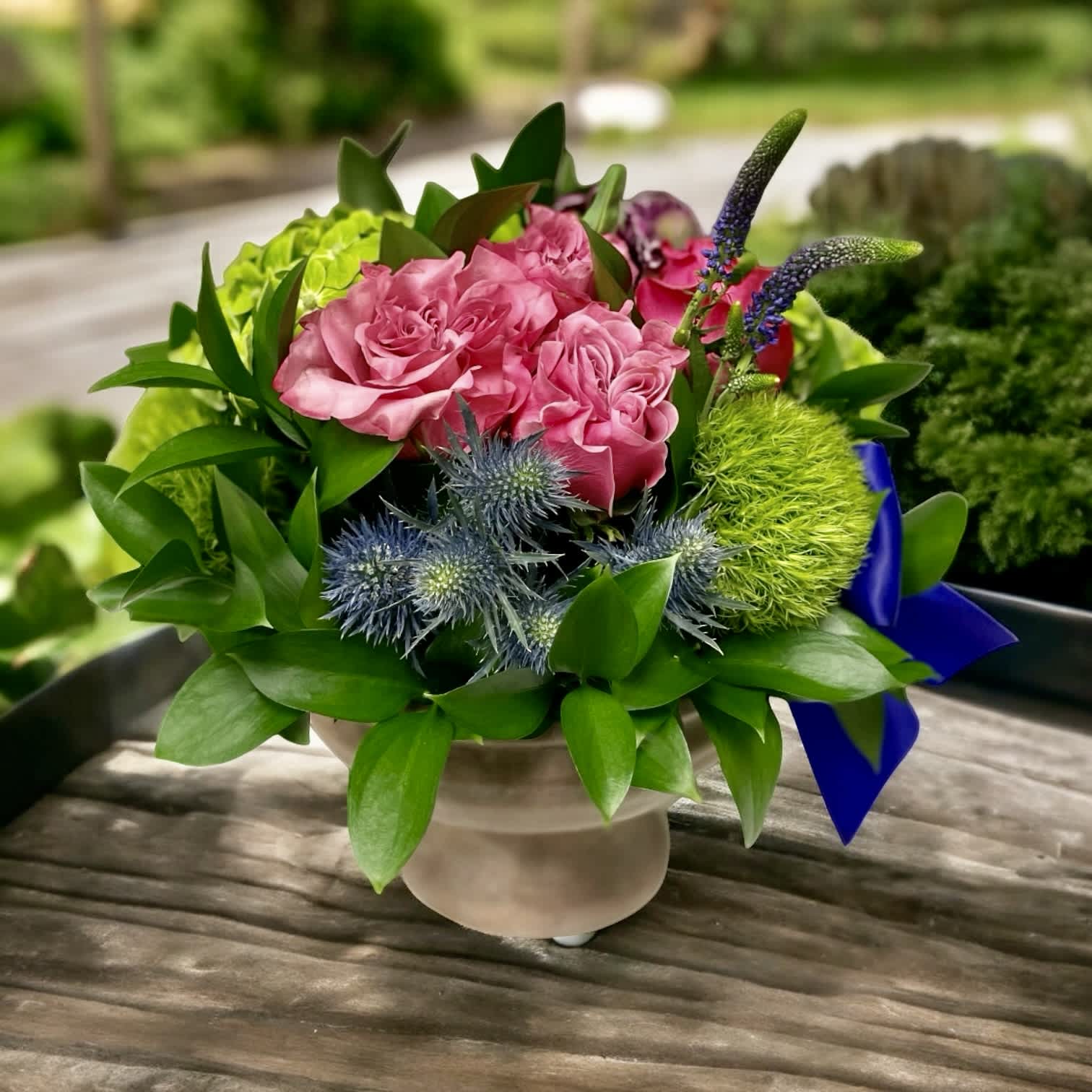 French Garden - French Garden – Garden-Style Floral Arrangement with Elegant Pedestal | Wilmington, Delaware Local Florist  Bring the charm of the French countryside to any space with the French Garden, a sophisticated floral arrangement designed by your trusted Wilmington, Delaware florist. This lush garden-style design features fresh green hydrangea, romantic pink garden roses, and striking blue thistle. Artfully arranged in a classic stone pedestal container and finished with a rich purple satin ribbon, French Garden exudes timeless beauty and elegance.  Perfect for birthdays, anniversaries, sympathy, or as a unique gift to brighten any room. Available for same-day flower delivery in Wilmington, DE and nearby areas. Count on our local flower shop for premium blooms, creative designs, and exceptional service.