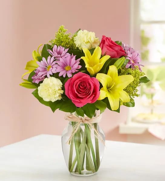 Bouquet of pink roses, yellow lilies, and purple daisies in a glass vase