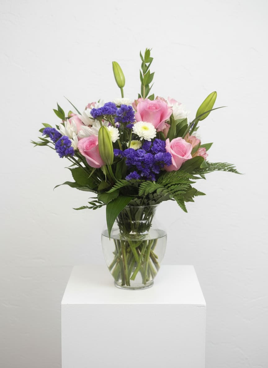 Blushing Bouquet  - Soft pink roses softened by crisp white daisy-like blooms and pops of vivid purple filler create a cheerful, romantic birthday bouquet that feels like spring in a clear glass vase. The unopened lily buds rising above the rounded mix give the arrangement a graceful, slightly taller profile-perfect for celebrating a birthday, saying "I'm thinking of you," or brightening someone's day.  This Blushing Bouquet features roses, lilies (with buds for a gentle reveal), daisy-style flowers, and seasonal filler arranged in a simple clear glass vase so the stems and fresh greenery are part of the visual charm. The palette of pink, white, purple, and green reads fresh and uplifting; the layered textures-from velvety rose petals to the lacy purple clusters-make the bouquet feel both lush and lively.  Choose this arrangement when you want the recipient to feel seen and celebrated: it's the kind of bouquet that brings a smile at a kitchen table, on a bedside table, or as a centerpiece for a small spring gathering. Vase included for easy display; lilies will open over a few days-trim stems and change the water regularly to keep blooms looking their best.