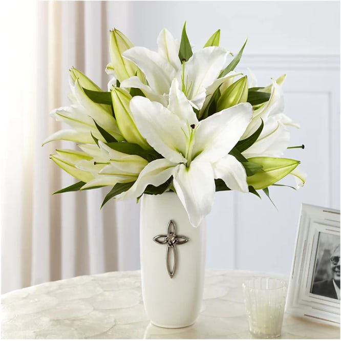 White lilies arranged in a tall white vase with a cross emblem