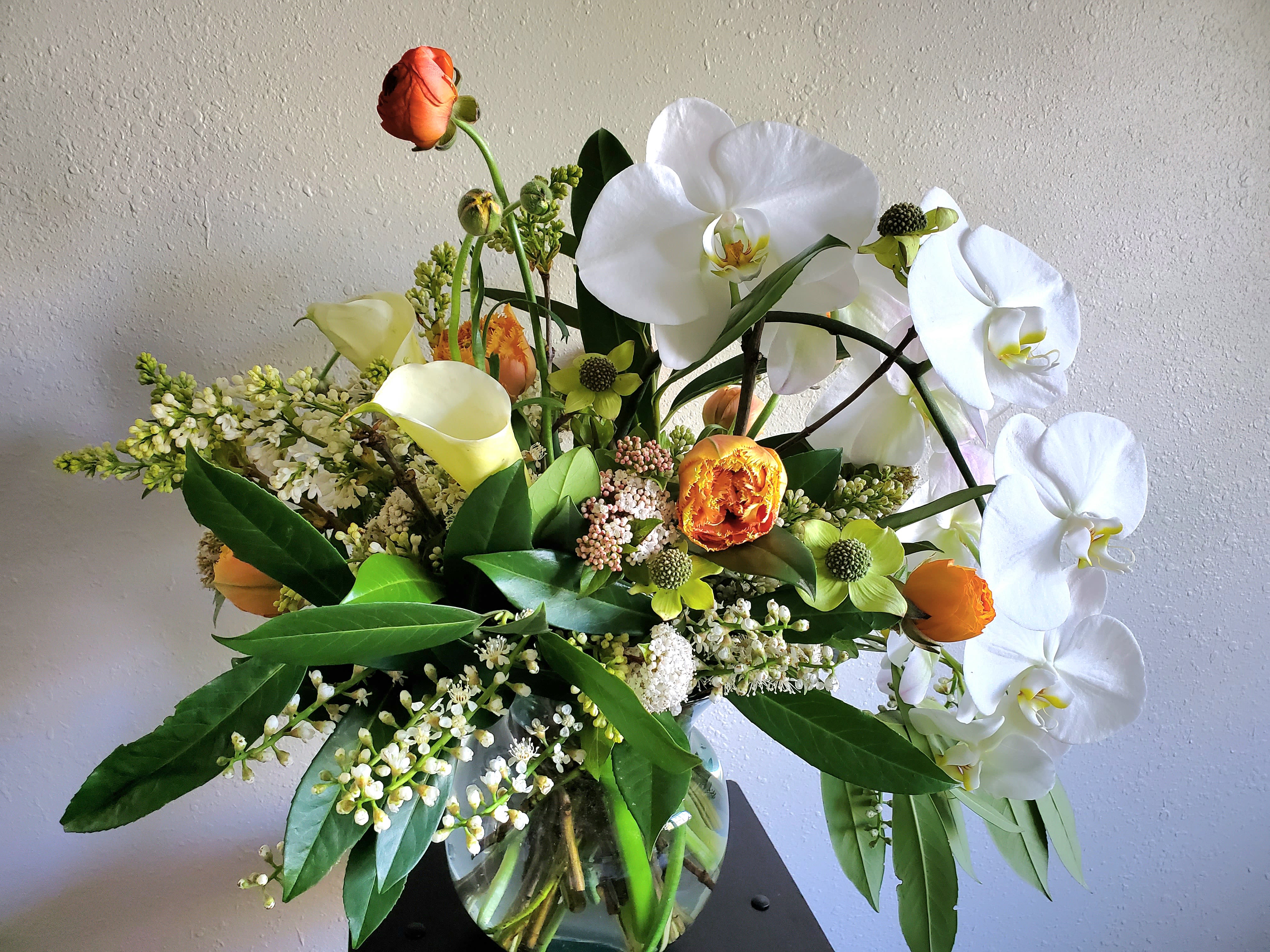 Arrangement of white orchids, yellow calla lilies, and orange ranunculus in a clear glass vase