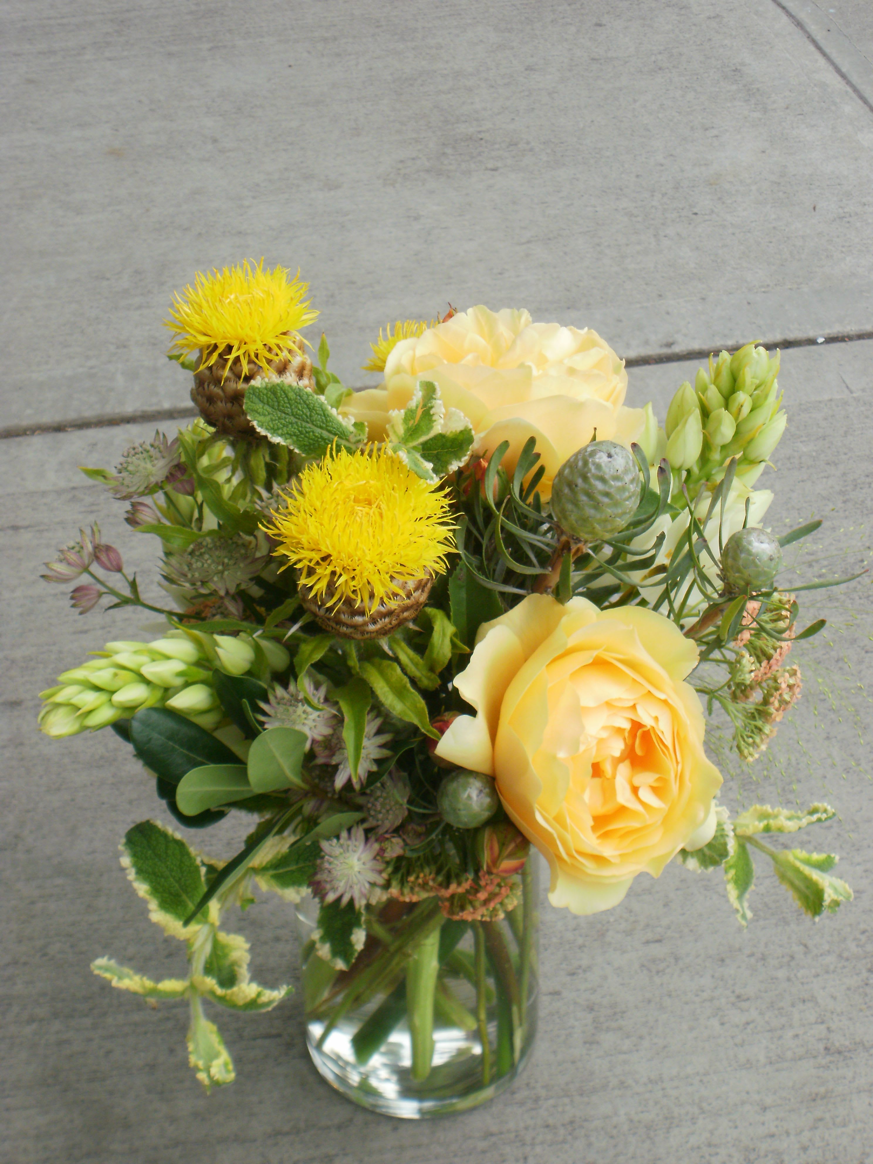 Yellow roses and spiky yellow blooms with green pods in a clear glass vase