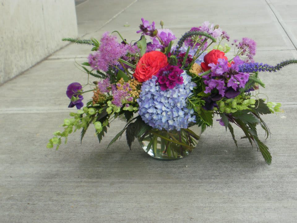 Low glass vase of mixed purple, blue, and red flowers arranged in a wide garden-style centerpiece