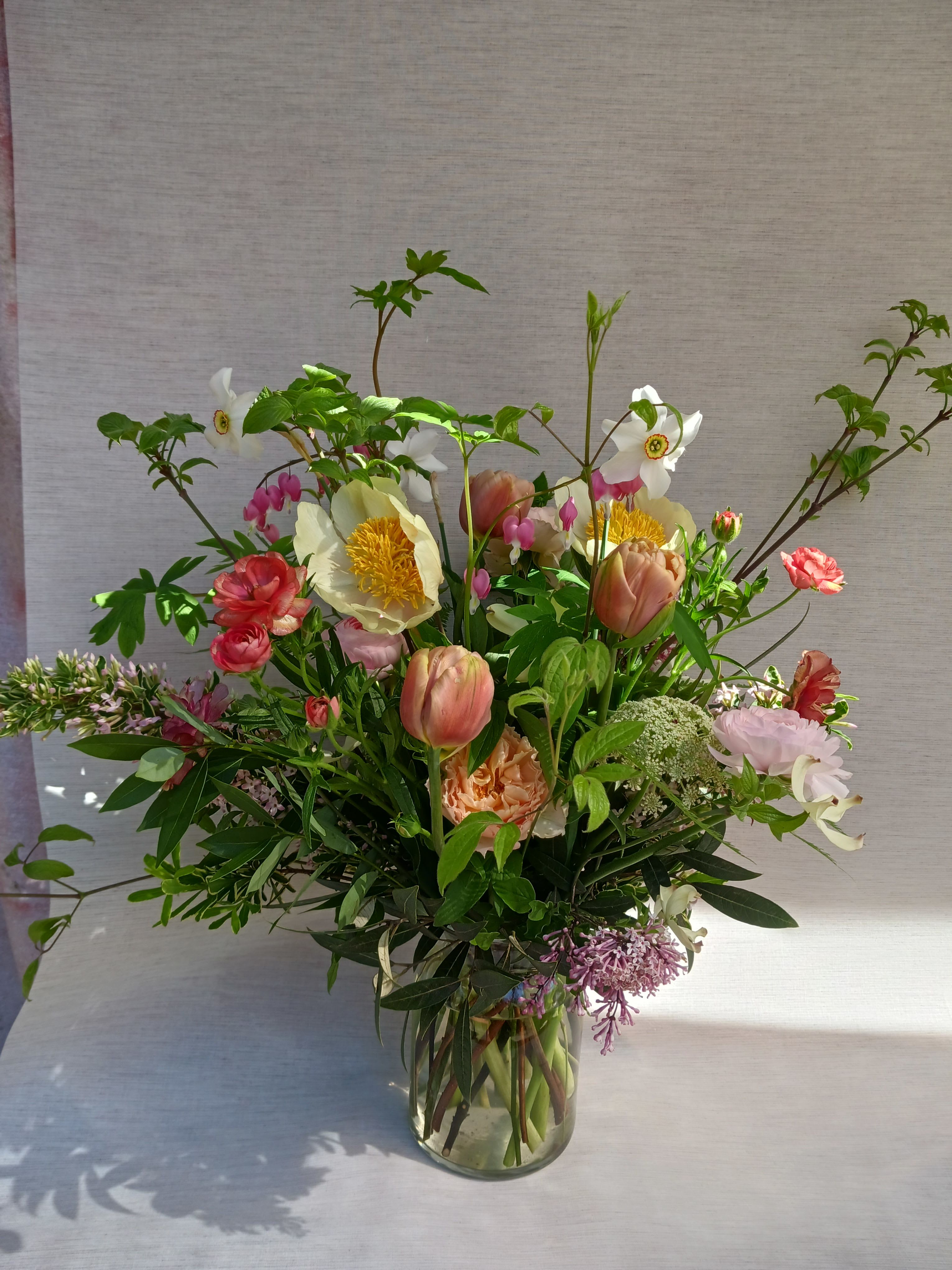 Tall garden-style arrangement of pink, peach, and white flowers with branches in a clear glass vase.