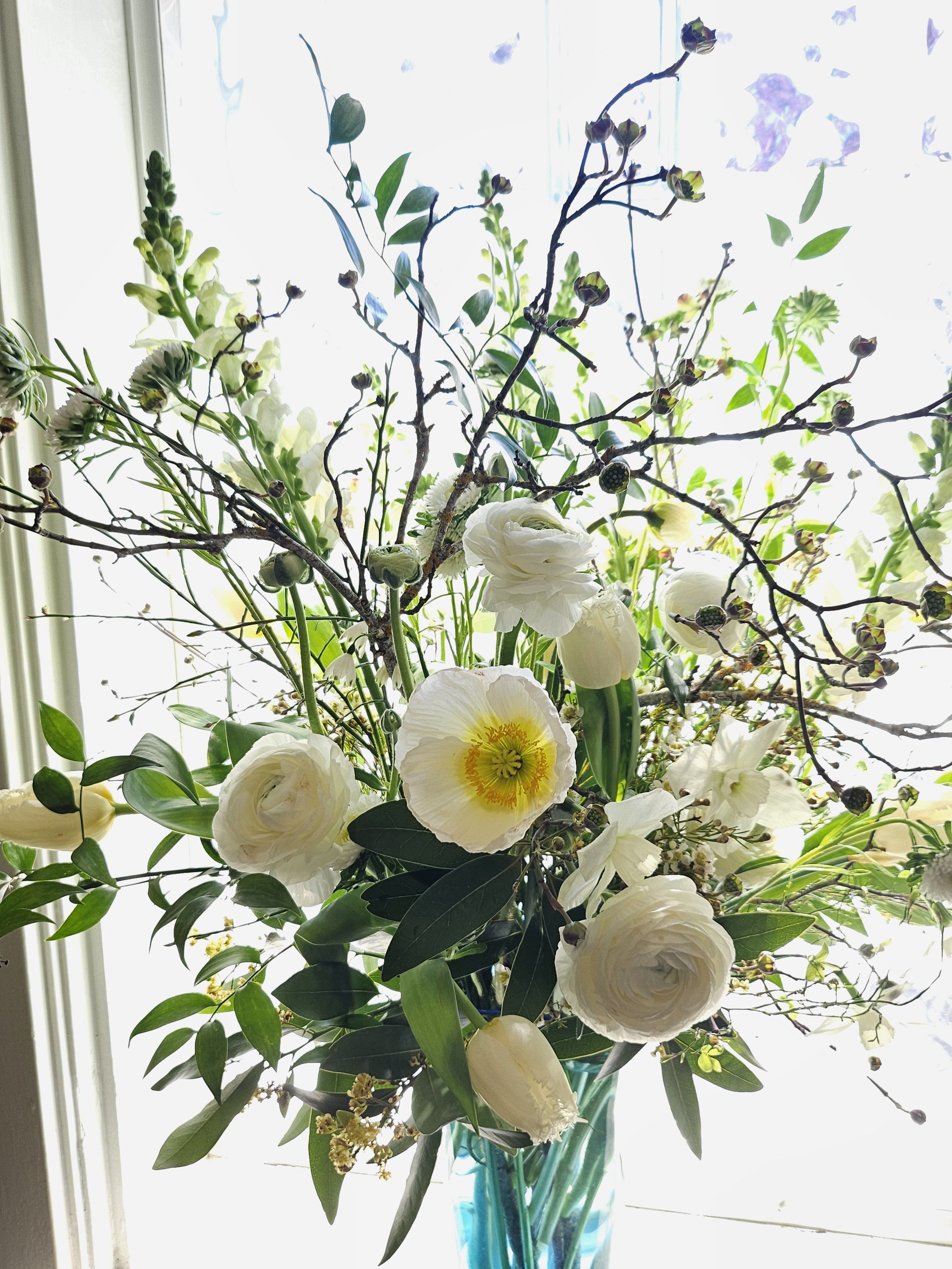 Tall arrangement of white flowers and twiggy branches in a blue glass vase by a bright window