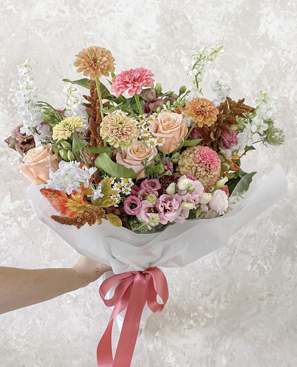 Handheld bouquet of peach, pink, and white flowers wrapped in white paper with a pink ribbon.