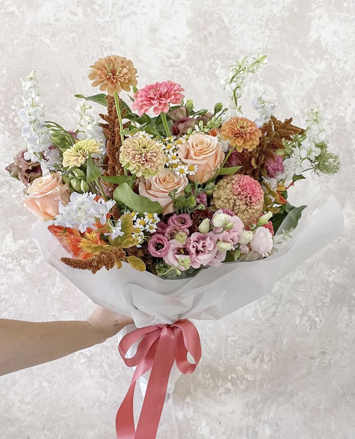 Handheld bouquet of peach, pink, and white flowers wrapped in white paper with a pink ribbon.