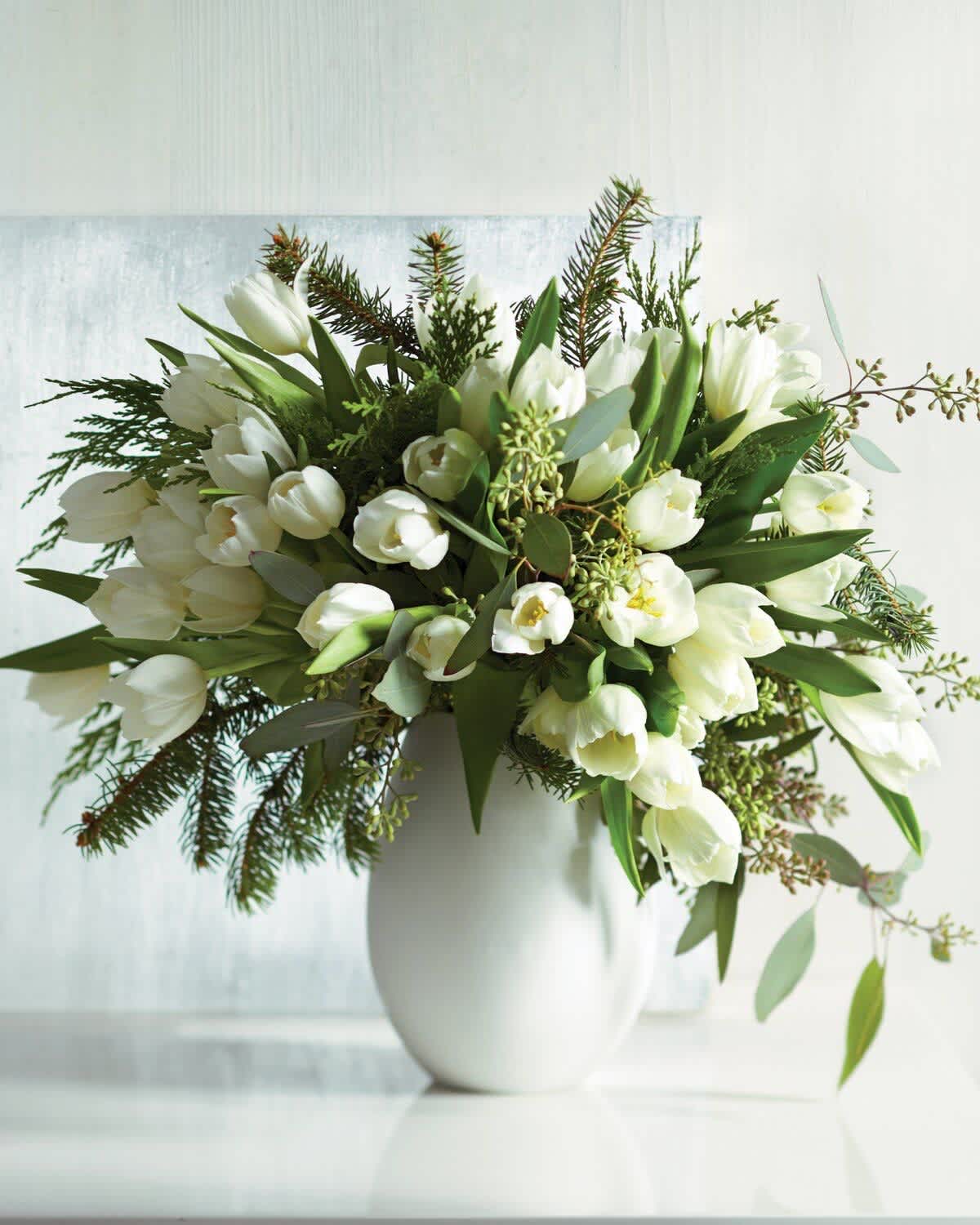 White tulips arranged in a white vase with evergreen foliage