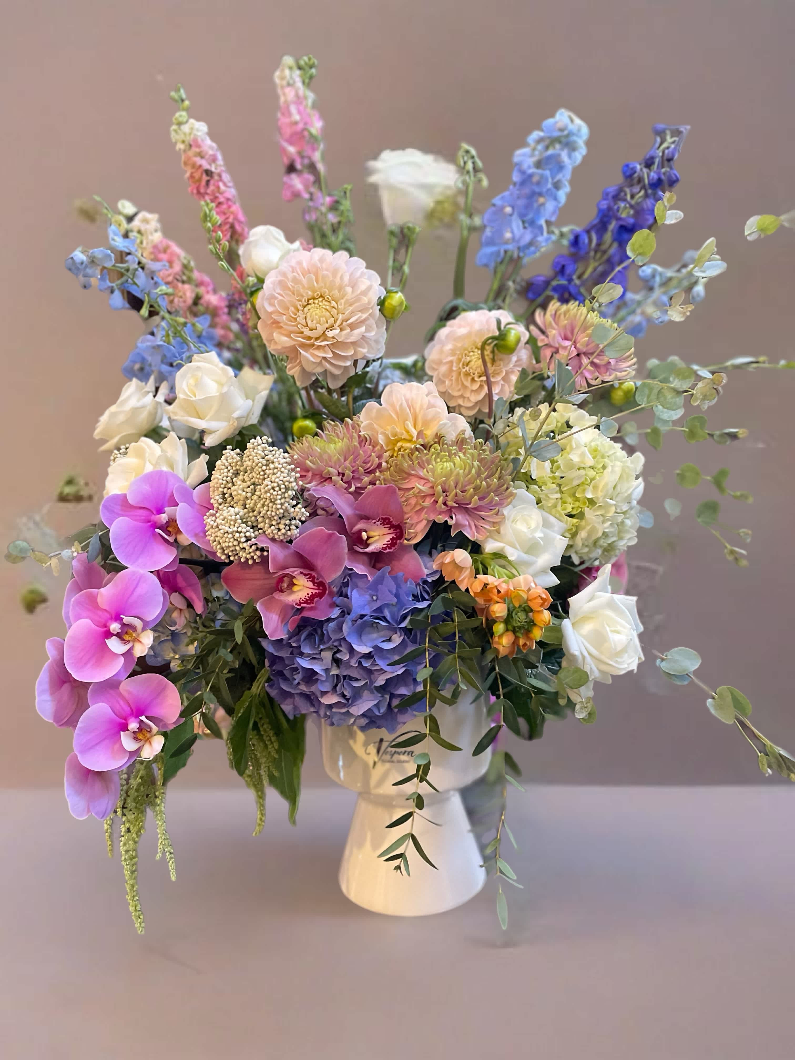 Mixed pastel bouquet in a white vase with orchids, roses, hydrangea, and tall blue blooms