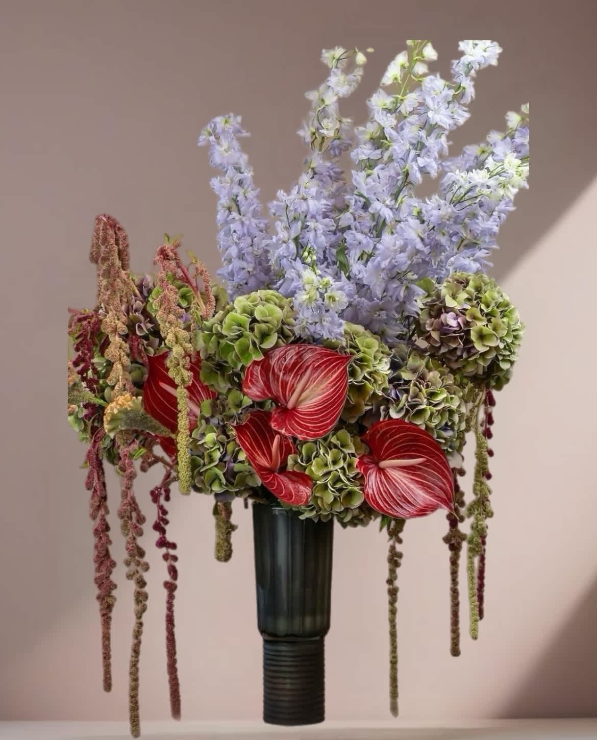 Tall arrangement with red anthuriums, green hydrangeas, and pale lavender flowers in a dark vase.