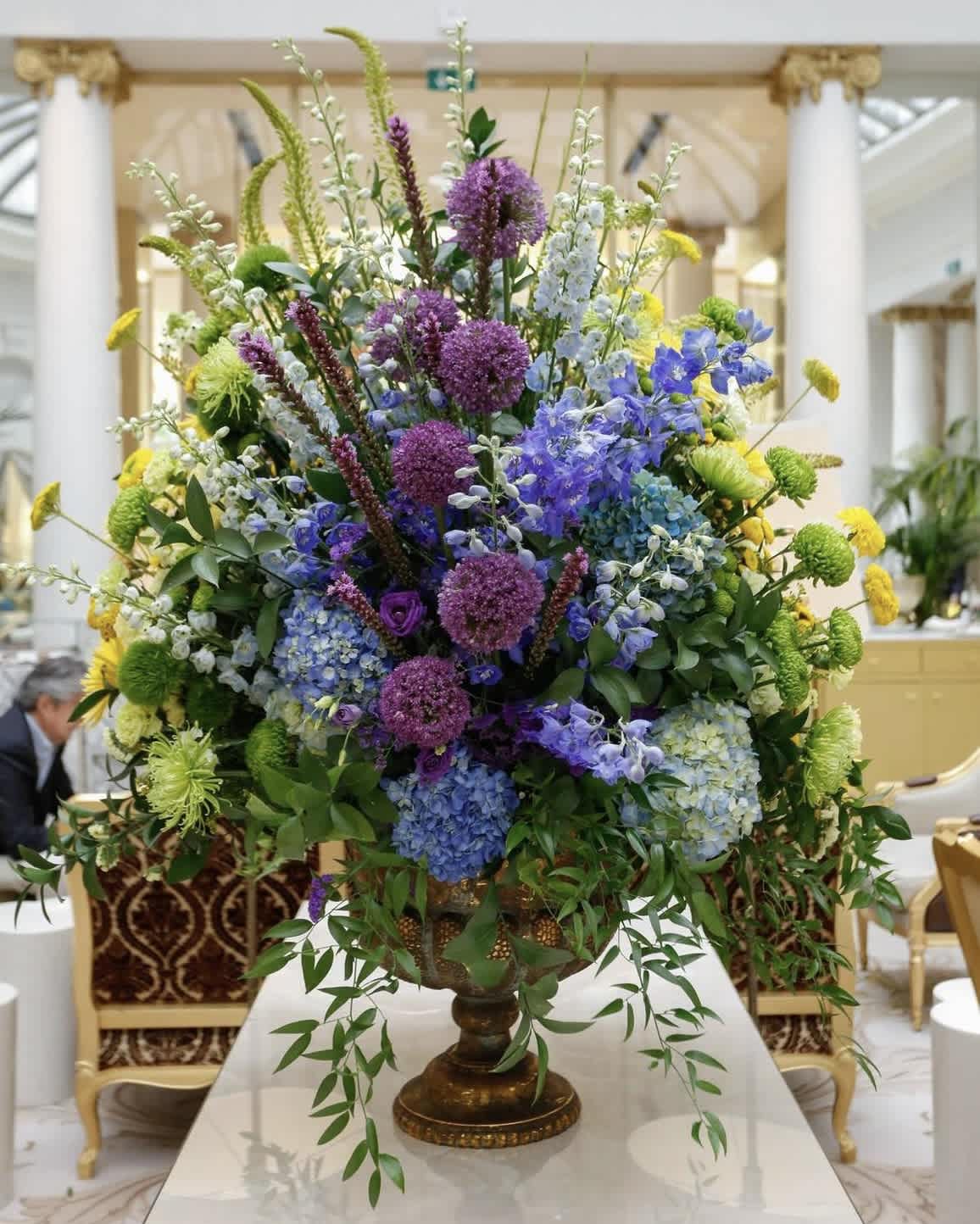 Large mixed floral centerpiece in a pedestal vase with purple, blue, yellow, and green blooms