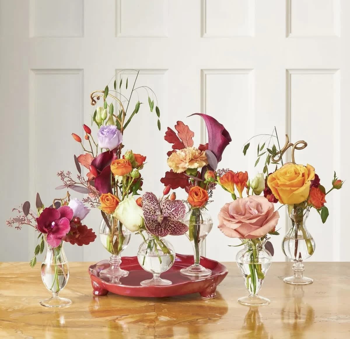 Five small floral arrangements in clear glass vases on a red tray