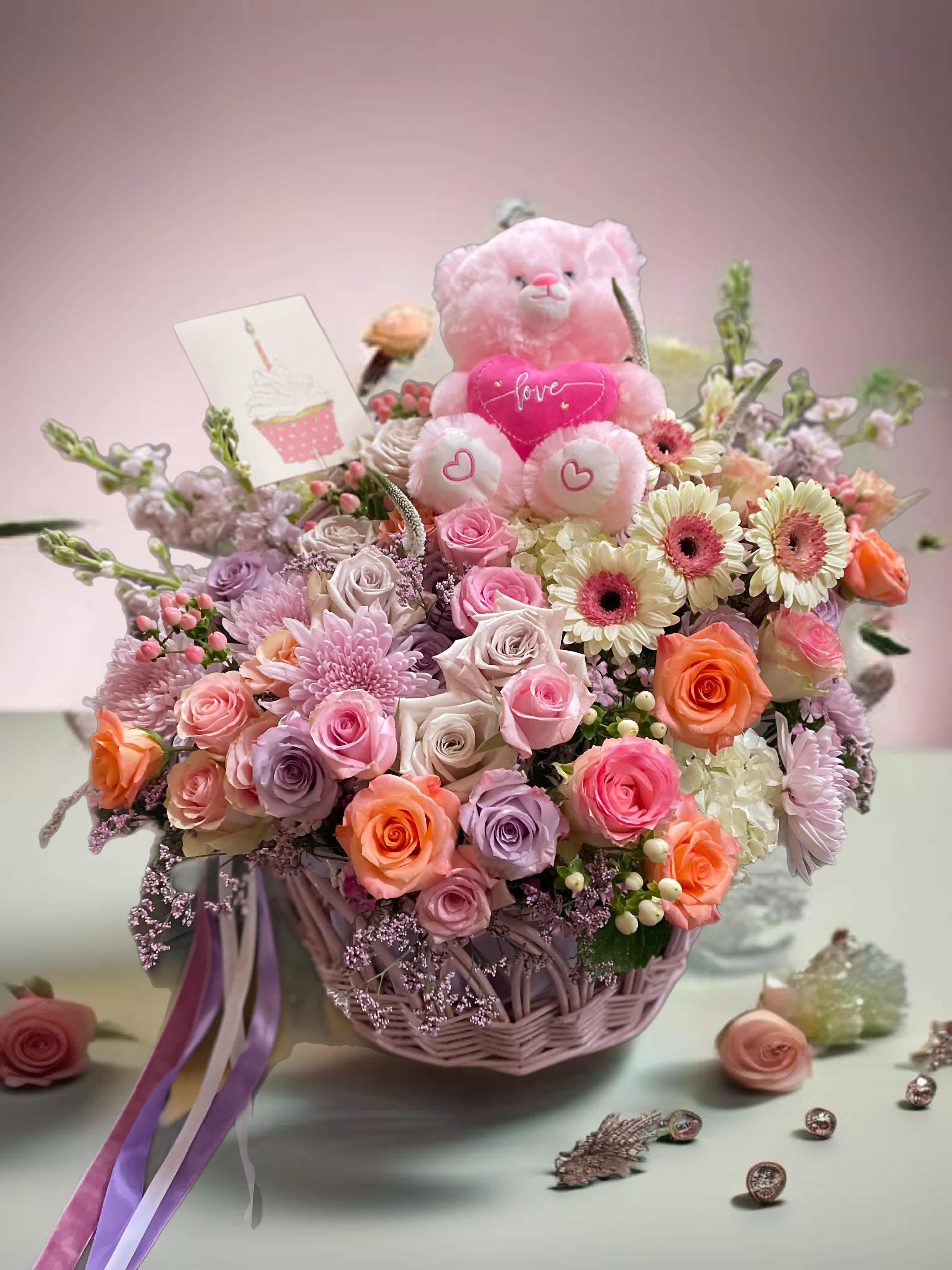 Pink teddy bear in a basket of roses and daisies with a love card
