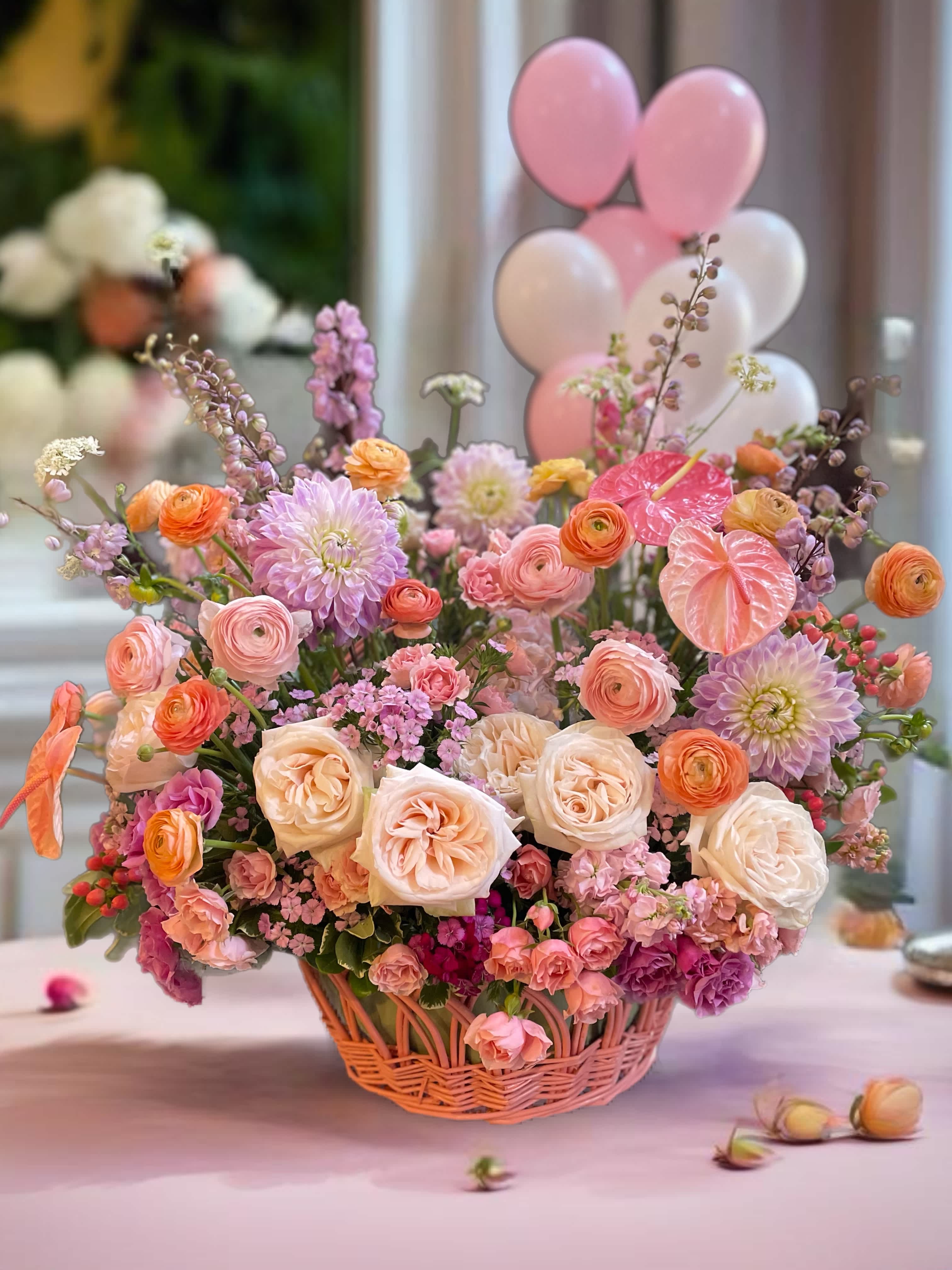 Pastel flower arrangement in a wicker basket with pink balloons behind it