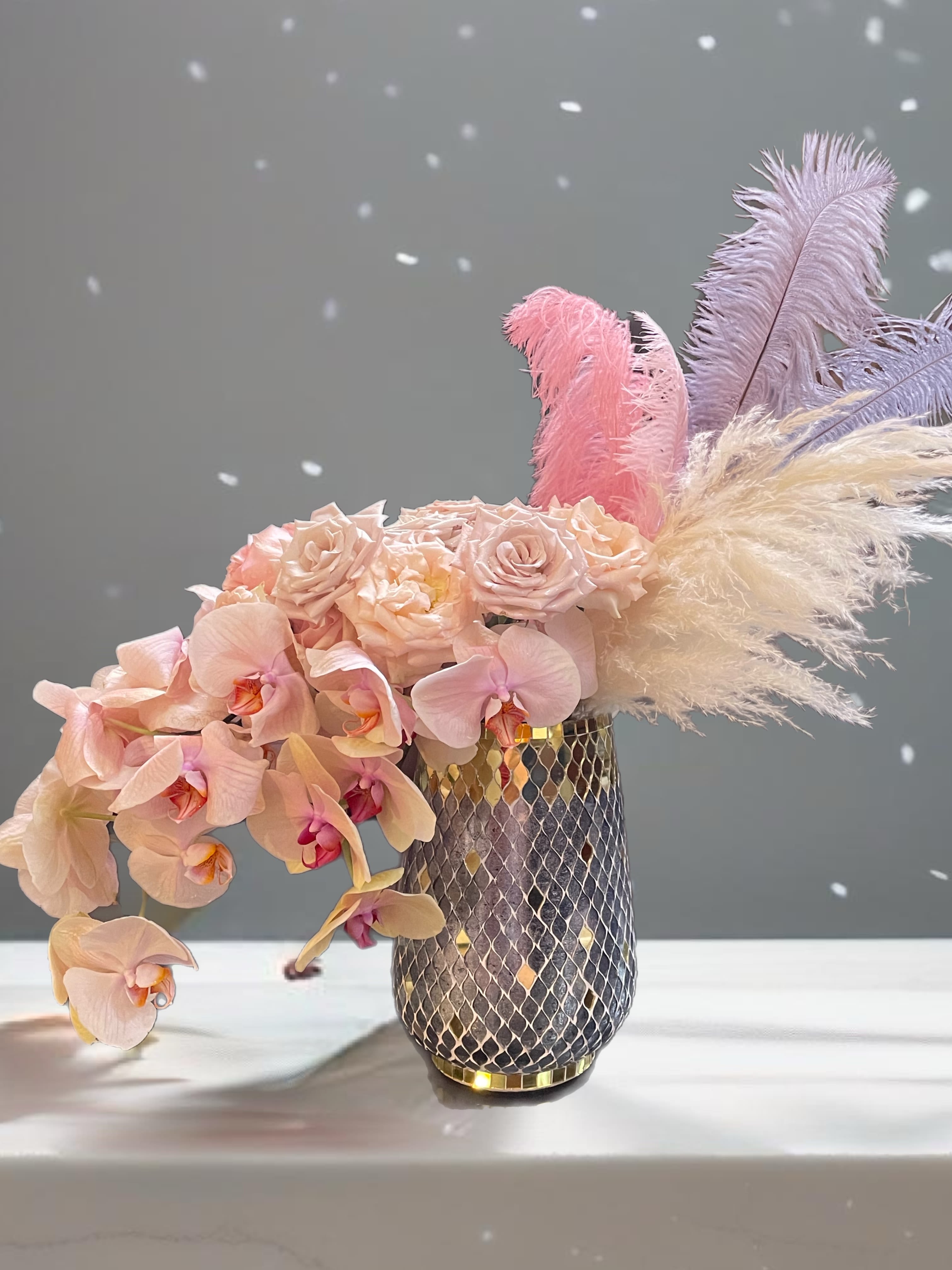 Pink orchids and roses in a patterned vase with pastel feathers