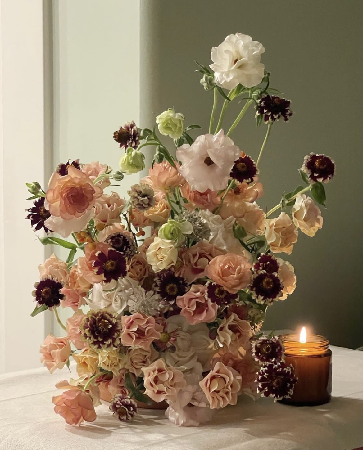 Tall bouquet of peach, blush, white, and burgundy flowers beside a lit candle