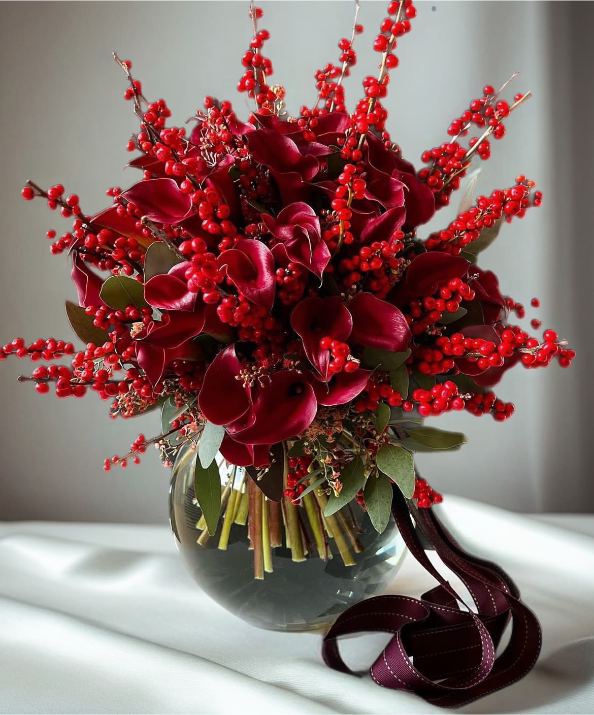 Deep red floral arrangement in a glass vase with berry branches and ribbon
