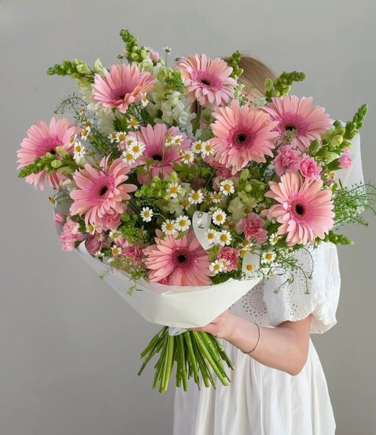 Simply gorgeous flowers  - Soft pink gerbera daisies take center stage in this large hand-tied bouquet, a cheerful birthday bouquet or everyday celebration arrangement that reads fresh and springlike. The mix of pink carnations and tall snapdragons gives soft height and texture while small white daisies with sunny yellow centers scatter like meadow flecks-she'll feel truly seen by the gentle, feminine palette.  This bouquet is gathered with long, visible stems and wrapped in crisp white paper, finished with a simple ribbon for a clean, modern presentation perfect for handing to a loved one or placing on a bedside table. Expect a generous size with abundant blooms and airy snapdragon spikes that add vertical movement and fragrance.  Ideal for birthdays, thank-you gestures, new baby visits, or "just because" moments, this arrangement reads joyful without being fussy-the kind of bouquet that brightens a room and the recipient's day. Care: trim stems and place in fresh water on arrival to extend vase life.