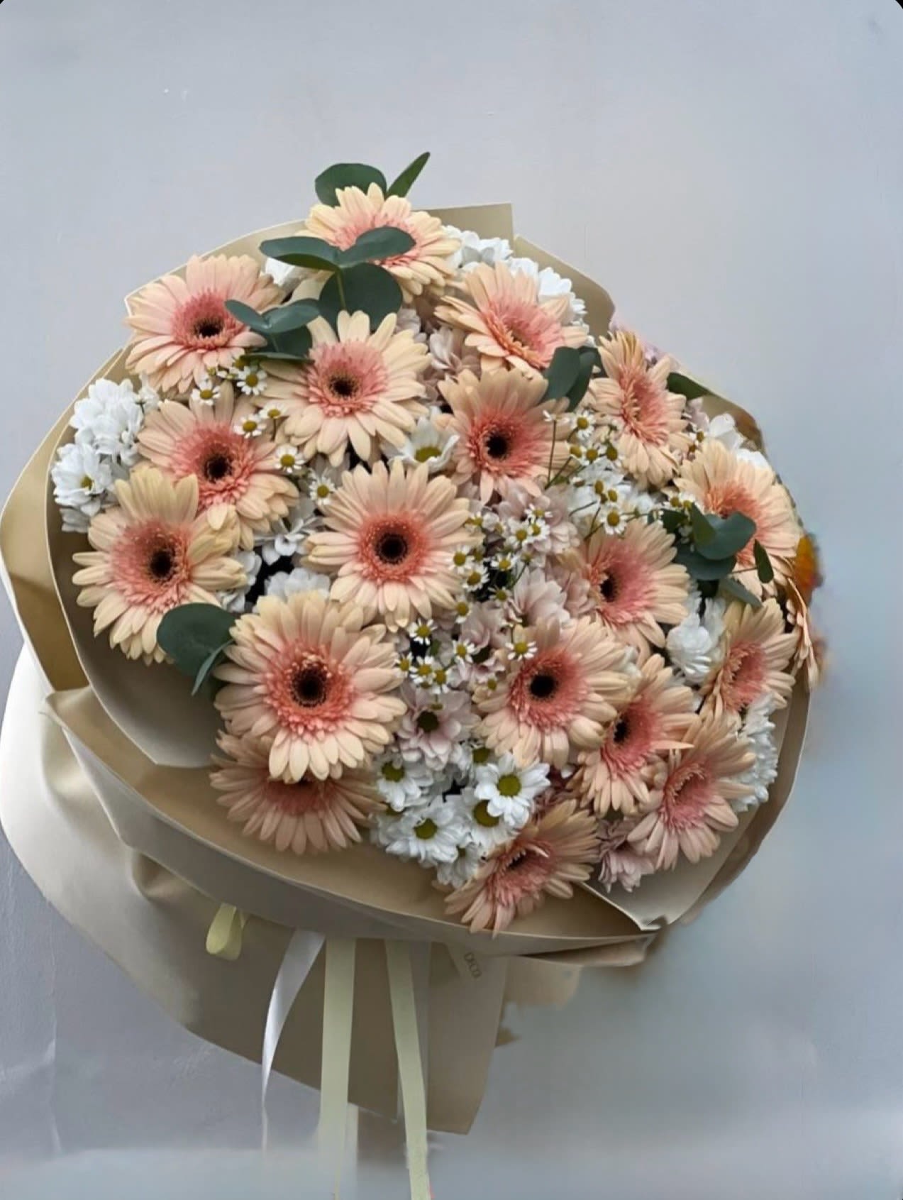 Happiest Days  - Soft peach gerbera daisies take center stage in this hand-tied flower arrangement, paired with delicate white daisies and full white chrysanthemums for a light, airy feel-perfect as a birthday bouquet, an anniversary surprise, a new baby gift, or a simple everyday gesture of care. The pastel peach centers against crisp white petals create a gentle, uplifting impression that will make the person you're thinking of feel warm and celebrated.  The blooms are gathered into a rounded, hand-tied shape and wrapped in layered beige paper, finished with long cream ribbons for a tidy, gift-ready presentation. Colors include peach, white, soft pink undertones, and fresh green foliage; textures range from the smooth gerbera faces to the feathery chrysanthemum petals and tiny daisy accents. Sized to sit prettily on a table or bedside, this bouquet arrives ready to display-no vase required-and is an effortless way to brighten someone's day.
