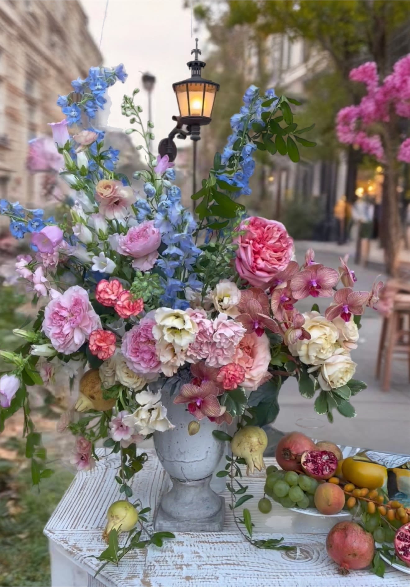 Bridgeton Spring Festival  - A lush, garden-style centerpiece that reads like a spring celebration-perfect as a spring gathering centerpiece, birthday arrangement, or an elegant everyday display. This large flower arrangement in a gray urn vase pairs tall blue delphinium-like spikes with layers of pink, cream, blush, and mixed-color blooms to create a romantic, celebratory presence that makes the person you're thinking of feel seen and cherished.  Built with roses, orchids, carnations, lisianthus, and complementary seasonal stems, the design is airy at the top and fully rounded at the base, with trailing greenery softening the edges. The composition sits in a substantial gray urn on the table, styled with a decorative fruit platter of pomegranates, grapes, pears, and mangoes for a bountiful, festive look-ideal for dressing a dining table or as the focal point of a party. Standard care: keep water fresh, trim stems every few days, and place out of direct heat to extend bloom life.