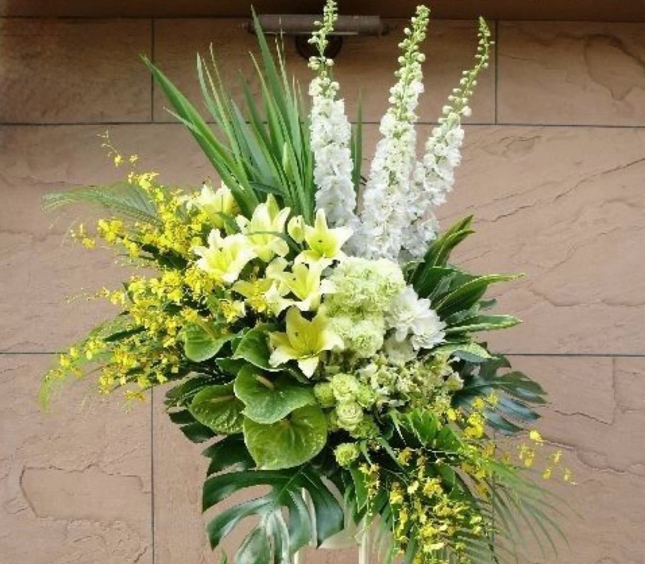 Podium stage arrangement  - A towering, ceremonial display built to command a stage-this podium stage arrangement is a formal sympathy or memorial piece designed to sit beside a lectern or on a raised platform. White lilies and orchid spikes create strong vertical lines while pale green hydrangea-like clusters and soft yellow filler blooms add a fresh, uplifting contrast; the overall effect reads elegant and slightly exotic, the kind of arrangement that helps the person you're honoring feel deeply respected and seen.