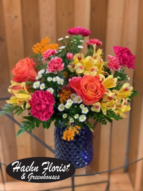 Bright Blooms Bouquet - A bold, sunlit mix that makes the person you're thinking of feel instantly celebrated - the Bright Blooms Bouquet is a mixed flower arrangement perfect for birthdays, get-well wishes, "just because" surprises, or a bright Labor Day centerpiece. Coral-pink roses and large bicolor sunflowers or yellow gerbera daisies take center stage, while hot pink carnations and orange roses add warm, energetic contrast. Mentioning occasions like Thank You, I'm Sorry, or Spring and Summer celebrations helps guide the moment this bouquet fits best without limiting its everyday appeal.  This design is available in several handsome vessels to suit the recipient's style: a dark blue diamond-patterned textured glass vase for a vintage, substantial look; a deep blue textured glass vase that makes colors pop; or a clear glass vase for a classic clustered-stem presentation. Purple filler blooms, airy white daisy-style flowers, yellow alstroemeria, green button-style blooms and a touch of succulent texture add visual interest, depth, and contrasting textures. The arrangement is full and rounded with varied heights for a casual, garden-fresh feel that will brighten a bedside table, desk, or dining room.