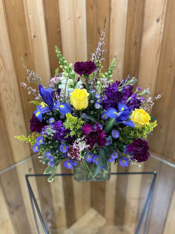 Mixed bouquet of yellow roses, blue irises, and purple flowers in a glass vase