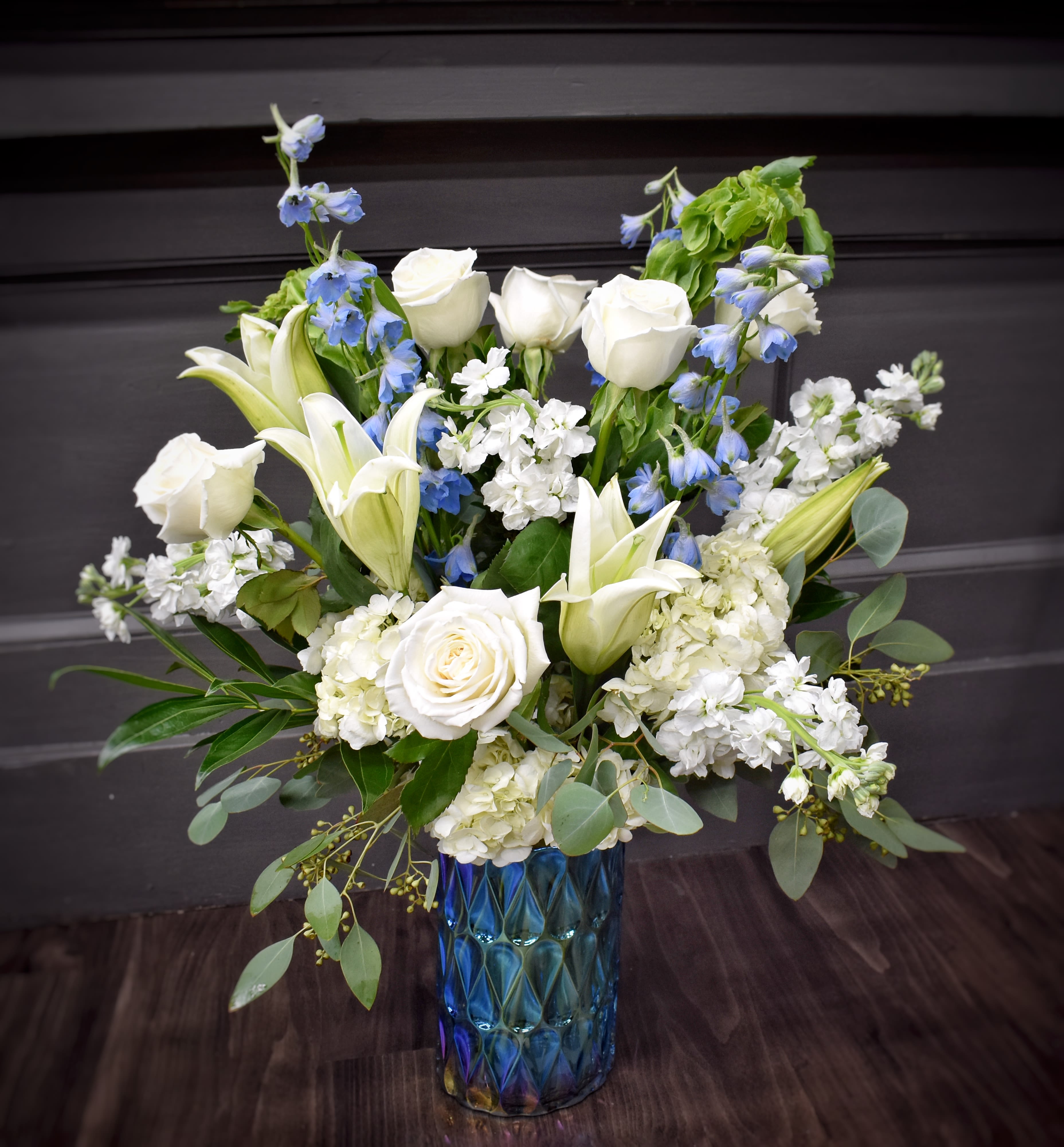 Blue Skies Premium - A serene wash of white and blue creates a calming first impression-perfect for a sympathy arrangement, a thoughtful thank-you, or a quiet celebration. This flower arrangement pairs classic white roses and lilies with airy blue delphinium-like spikes, giving the bouquet a graceful vertical movement that feels both elegant and gently uplifting. The overall effect is refined and tranquil, the kind of gift that helps someone feel seen and comforted.  The composition is full and upright, built around clusters of white stock and hydrangea for soft volume, with soft green foliage and eucalyptus-like accents fanning around a sleek blue glass vase. Textures range from the smooth, velvety rose petals to the ruffled hydrangea and feathery delphinium, creating visual depth while staying within a fresh white, blue, and green palette. The blue glass vase grounds the arrangement and makes a subtle statement on mantels, desks, or bedside tables.  Ideal for the person you want to soothe, honor, or quietly celebrate, this arrangement reads as both formal enough for memorial settings and warm enough for everyday gifting. 