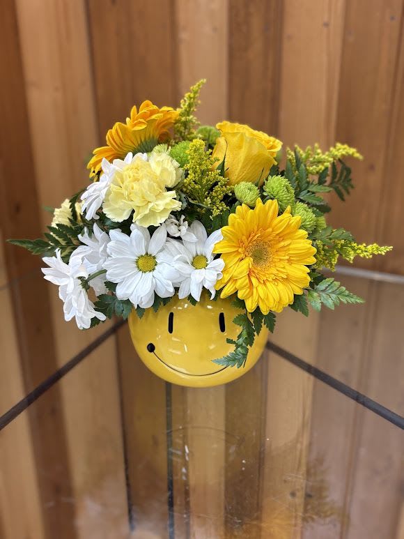 Yellow and white daisies, roses, and carnations in a yellow smiley-face ceramic vase.