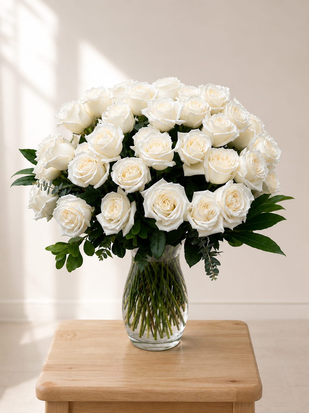 The Royal Rose Bouquet  - A cloudlike cluster of pure white roses creates an immediate impression of calm and refinement-perfect as a wedding bouquet centerpiece, a sympathy arrangement, or a graceful everyday gift. The Royal Rose Bouquet's rounded silhouette and serene white palette say elegance and respect, the kind of arrangement that makes the person you're thinking of feel comforted and truly honored.  Tightly gathered, full-layered white rose blooms sit low in a clear glass vase, with deep green foliage framing the base and peeking between petals for striking contrast. The compact, formal shape reads clean and restrained from every angle, delivering a luxe, photographed-ready look without being fussy.  Made to convey heartfelt sentiment for ceremonies, memorials, anniversaries, or to simply let someone know they're seen, this bouquet arrives ready to display-no extra styling needed. Keep stems in fresh water and recut if desired to extend vase life; sizing and exact stem counts may vary by availability to ensure each arrangement looks as full and flawless as pictured.
