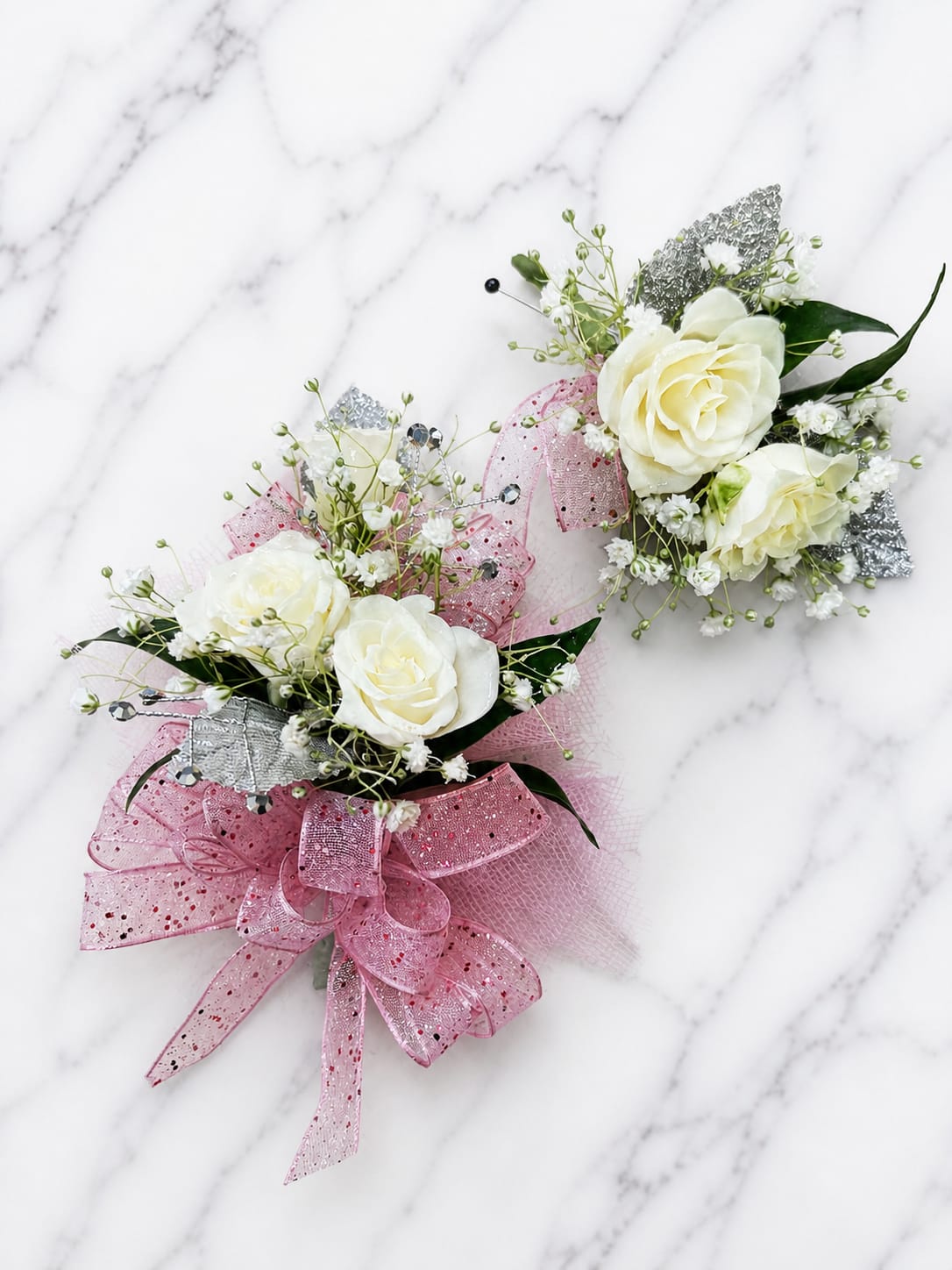 Pink Stardust Set - ALL BOUTONNIÈRES INCLUDE A MAGNETIC ATTACHMENT. MAGNET WARNING: IF THE WEARER HAS A PACEMAKER, DEFIBRILLATOR, INSULIN PUMP, HEARING DEVICE, OR OTHER MEDICAL EQUIPMENT, PLEASE REQUEST A TRADITIONAL PIN ATTACHMENT WHEN ORDERING.  Pink Stardust brings classic elegance and sparkle to prom, homecoming, and formal school dances. This matching corsage and boutonnière set features white spray roses accented with coordinating flowers, metallic leaves, ribbon, rhinestone accents, and fresh greenery for a timeless look that complements a wide variety of dress and tuxedo colors.  Each set includes a matching boutonnière featuring two white spray roses designed to coordinate with the corsage. Choose from Standard, Deluxe, or Premium options based on the number of roses in the corsage. Standard features 3 spray roses, Deluxe features 4 spray roses, and Premium features 5 spray roses for a fuller presentation.  Accent flowers and decorative elements may vary based on availability while maintaining the overall style, color palette, and value of the design. Each piece is handcrafted, and no two designs are alike for a local dance.