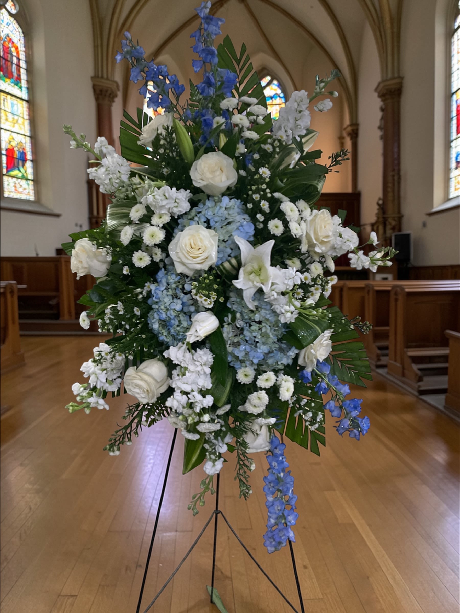 Sincerely Heartfelt Spray on Easel - Sincerely Heartfelt is a graceful standing spray designed to offer comfort, peace, and a heartfelt expression of sympathy. Featuring white roses, lilies, stock, soft blue hydrangea, and cascading blue delphinium, this elegant tribute combines timeless beauty with gentle color and natural movement. The calming palette of blue and white symbolizes peace, faith, remembrance, and enduring love, making it a meaningful choice for funeral services, memorial services, celebrations of life, and church services.  Designed with balance and flowing lines, Sincerely Heartfelt creates a beautiful presentation while allowing each flower to be appreciated individually. The combination of premium blooms, lush greenery, and delicate accents creates a tribute that feels uplifting, sincere, and deeply personal.  Whether honoring a parent, spouse, grandparent, sibling, friend, or cherished loved one, this standing spray serves as a lasting expression of love, respect, gratitude, and remembrance. Its classic design makes it appropriate for both traditional and contemporary services.  Available through Aspen Tower Florist for services at Weigel Funeral Home, Cook Rosenberger Funeral Home, and Neal’s Funeral Home, serving families in Batesville, Sunman, and Osgood.