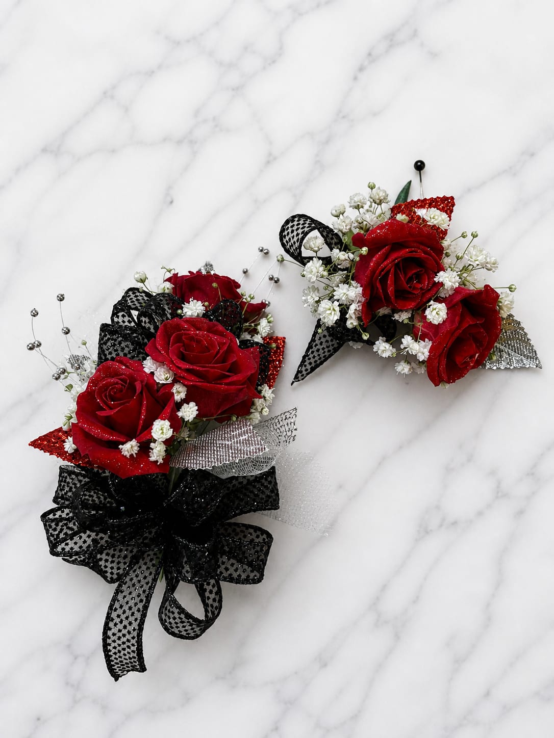 Ruby Roses Formal Set - MAGNETIC BOUTONNIERE WARNING: CONTAINS STRONG MAGNETS. DO NOT USE IF YOU HAVE A PACEMAKER, DEFIBRILLATOR, INSULIN PUMP, COCHLEAR IMPLANT, NEUROSTIMULATOR, OR OTHER IMPLANTED MEDICAL DEVICE. KEEP AWAY FROM ELECTRONICS, CREDIT CARDS, HOTEL KEY CARDS, AND CHILDREN.  Ruby Roses  Rich red roses, dramatic black ribbon, sparkling silver accents, and delicate white filler flowers come together in Ruby Roses, a bold corsage and boutonniere collection designed to make a statement. The striking combination of red, black, and silver creates a timeless formal look that is both elegant and unforgettable.  Standard includes a wrist corsage with 3 spray roses and a matching magnetic boutonniere with 2 roses. Deluxe includes 4 spray roses in the corsage. Premium includes 5 spray roses for additional fullness and flower coverage. All boutonnieres include 2 roses.  Perfect for prom flowers, wrist corsages, magnetic boutonnieres, corsage and boutonniere sets, homecoming flowers, dance flowers, and formal event flowers.  Because each corsage and boutonniere is individually designed, no two sets will be exactly alike. Ribbon, filler flowers, bracelets, decorative accents, and other design elements may vary based on availability. While individual details may differ, the overall color palette, roses and/or flowers, style, and appearance of the Ruby Roses collection will be maintained.
