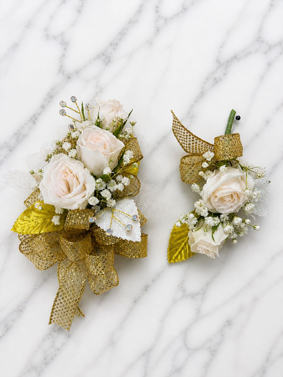 Gilded Blush Formal Set - MAGNETIC BOUTONNIERE WARNING: CONTAINS STRONG MAGNETS. DO NOT USE IF YOU HAVE A PACEMAKER, DEFIBRILLATOR, INSULIN PUMP, COCHLEAR IMPLANT, NEUROSTIMULATOR, OR OTHER IMPLANTED MEDICAL DEVICE. KEEP AWAY FROM ELECTRONICS, CREDIT CARDS, HOTEL KEY CARDS, AND CHILDREN.  Gilded Blush  Soft blush roses, shimmering gold ribbon, sparkling accents, and delicate filler flowers come together in Gilded Blush, an elegant corsage and boutonniere collection designed for prom, homecoming, dances, banquets, weddings, and formal events. The timeless combination of blush and gold creates a graceful, sophisticated look with just the right touch of sparkle.  Standard includes a wrist corsage with 3 spray roses and a matching magnetic boutonniere with 2 roses. Deluxe includes 4 spray roses in the corsage. Premium includes 5 spray roses for additional fullness and flower coverage. All boutonnieres include 2 roses.  Perfect for prom flowers, wrist corsages, magnetic boutonnieres, corsage and boutonniere sets, homecoming flowers, dance flowers, and formal event flowers.  Because each corsage and boutonniere is individually designed, no two sets will be exactly alike. Ribbon, filler flowers, bracelets, decorative accents, and other design elements may vary based on availability. While individual details may differ, the overall color palette, roses and/or flowers, style, and appearance of the Gilded Blush collection will be maintained.